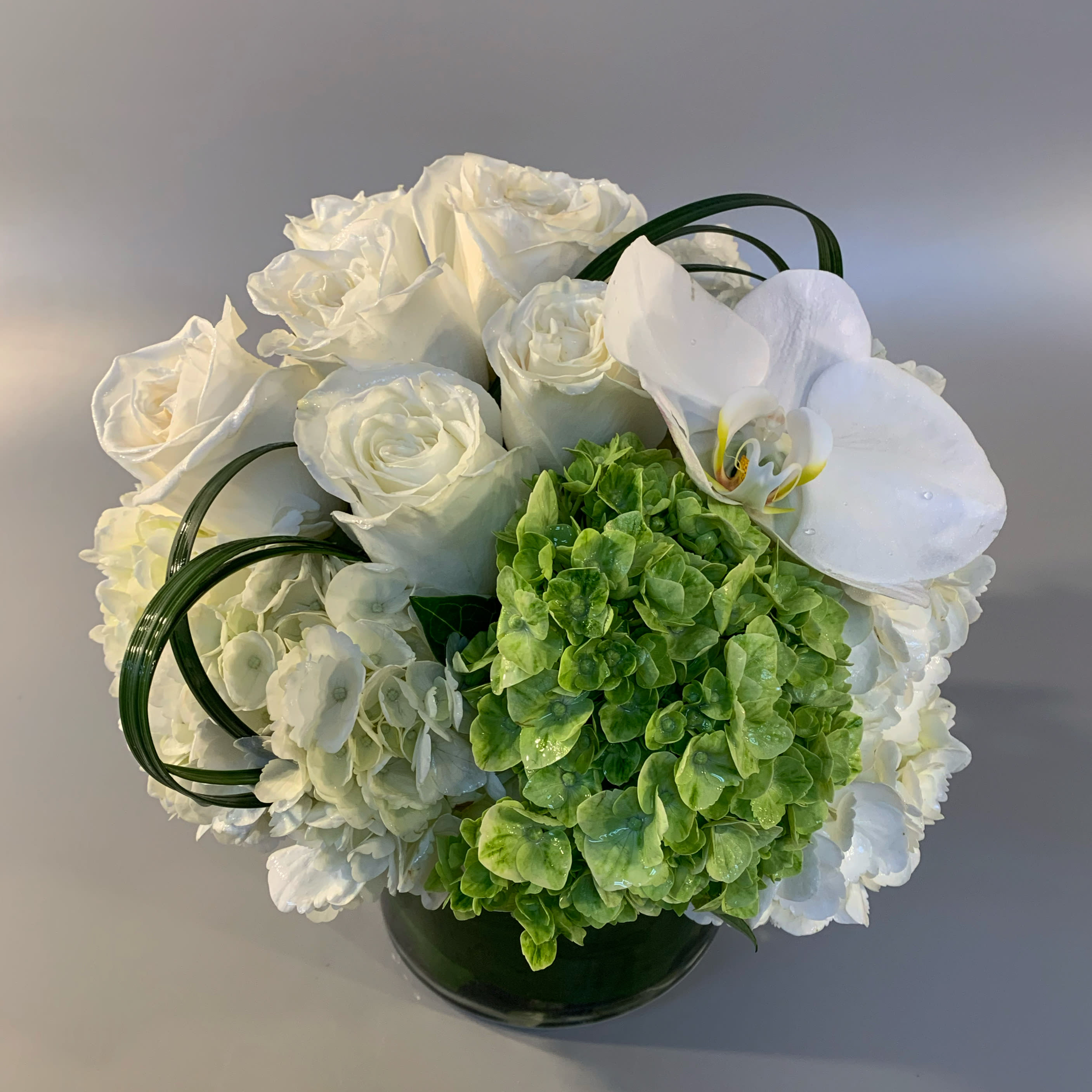 FRESH - A soft and sculptural hand tied arrangement designed with subtle shades of white and green hydrangea, orchid blossoms, sculpted grasses, and roses. Placed in a white glass vessel.