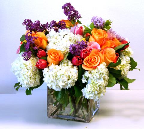 Low mixed arrangement of white hydrangeas, orange roses, and purple accents in a clear glass cube vase