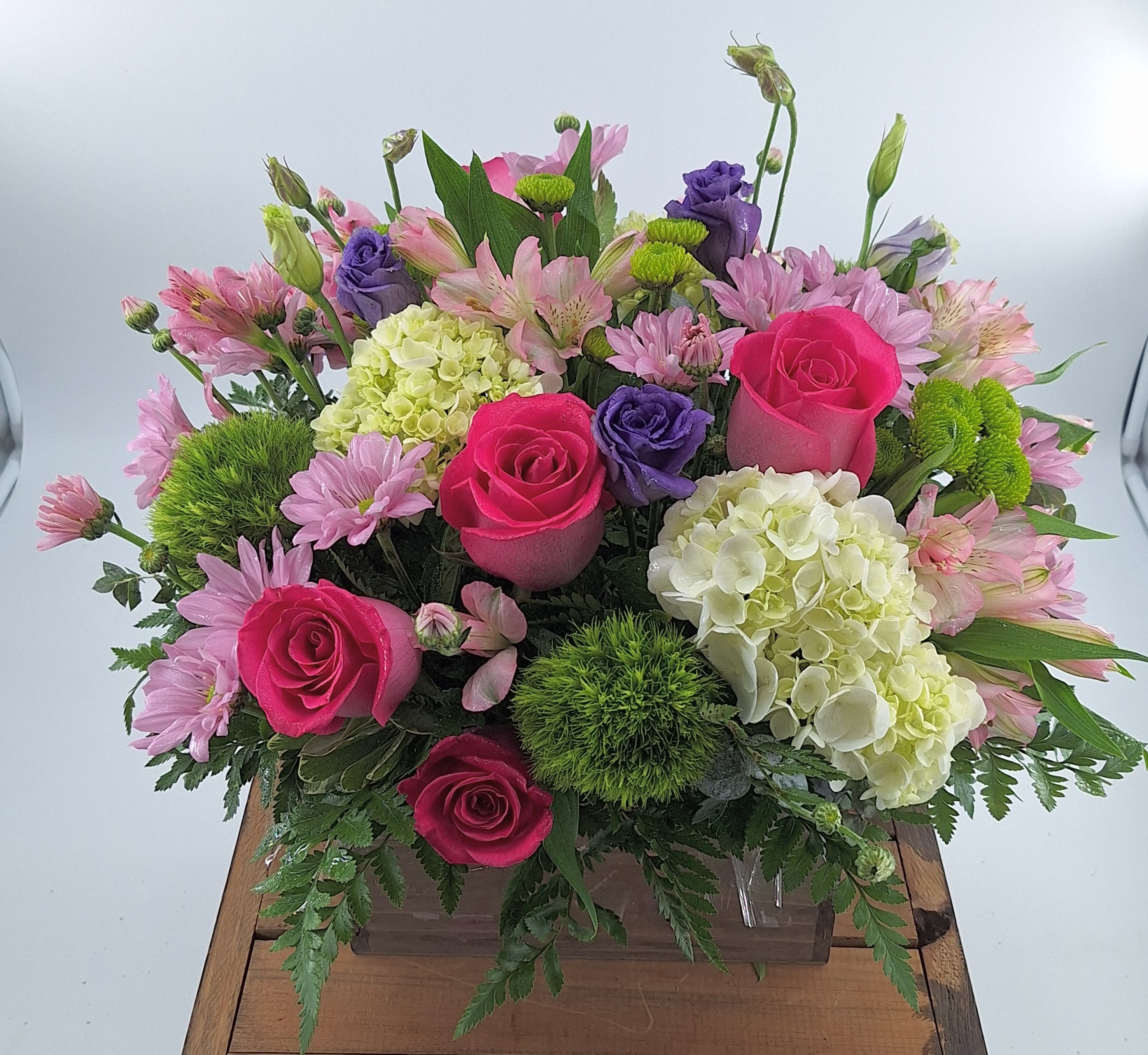 Mixed arrangement of hot pink roses, white hydrangeas, and purple blooms in a clear rectangular vase