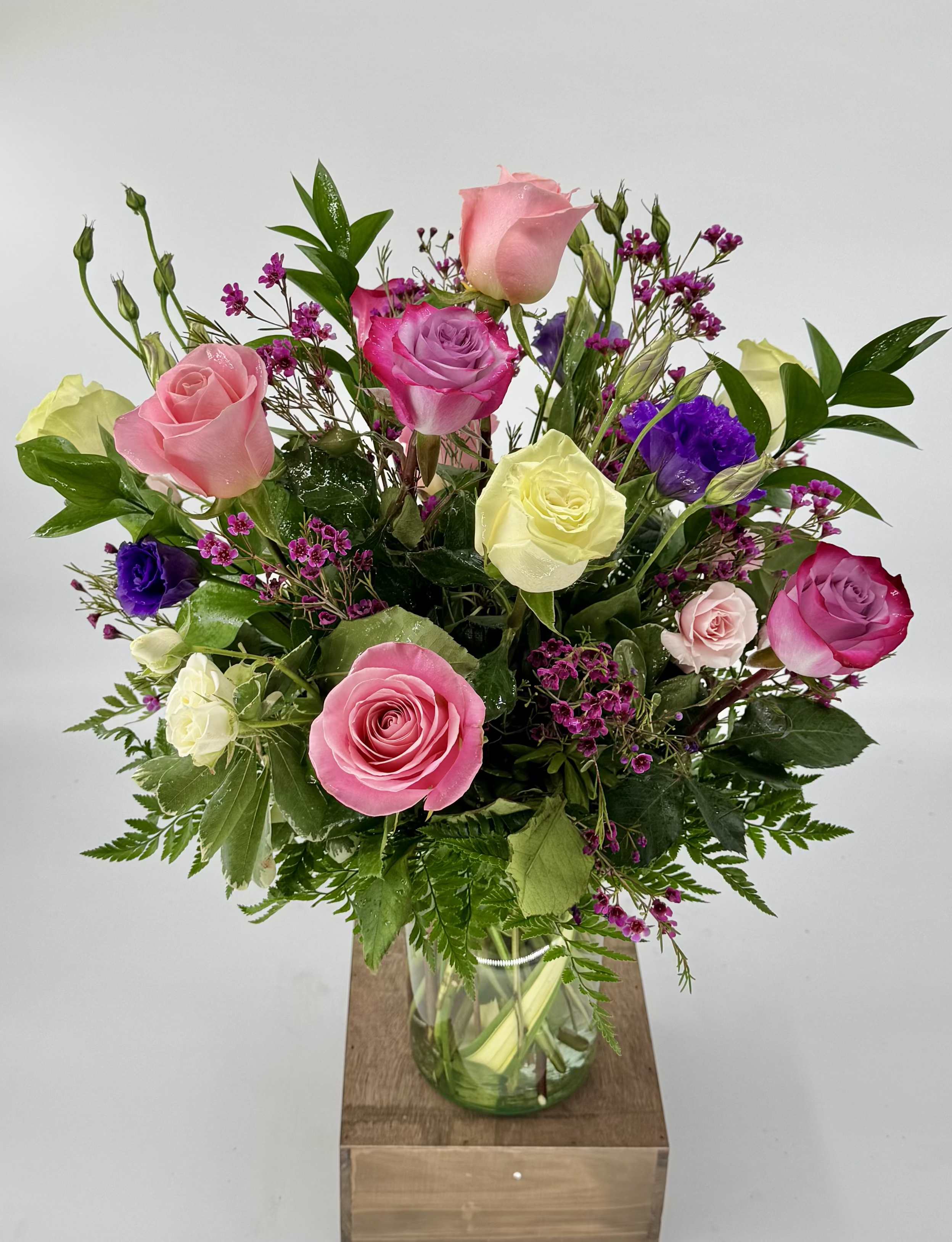 Mixed arrangement of pink, cream, and purple roses with small fuchsia blooms in a clear glass vase.