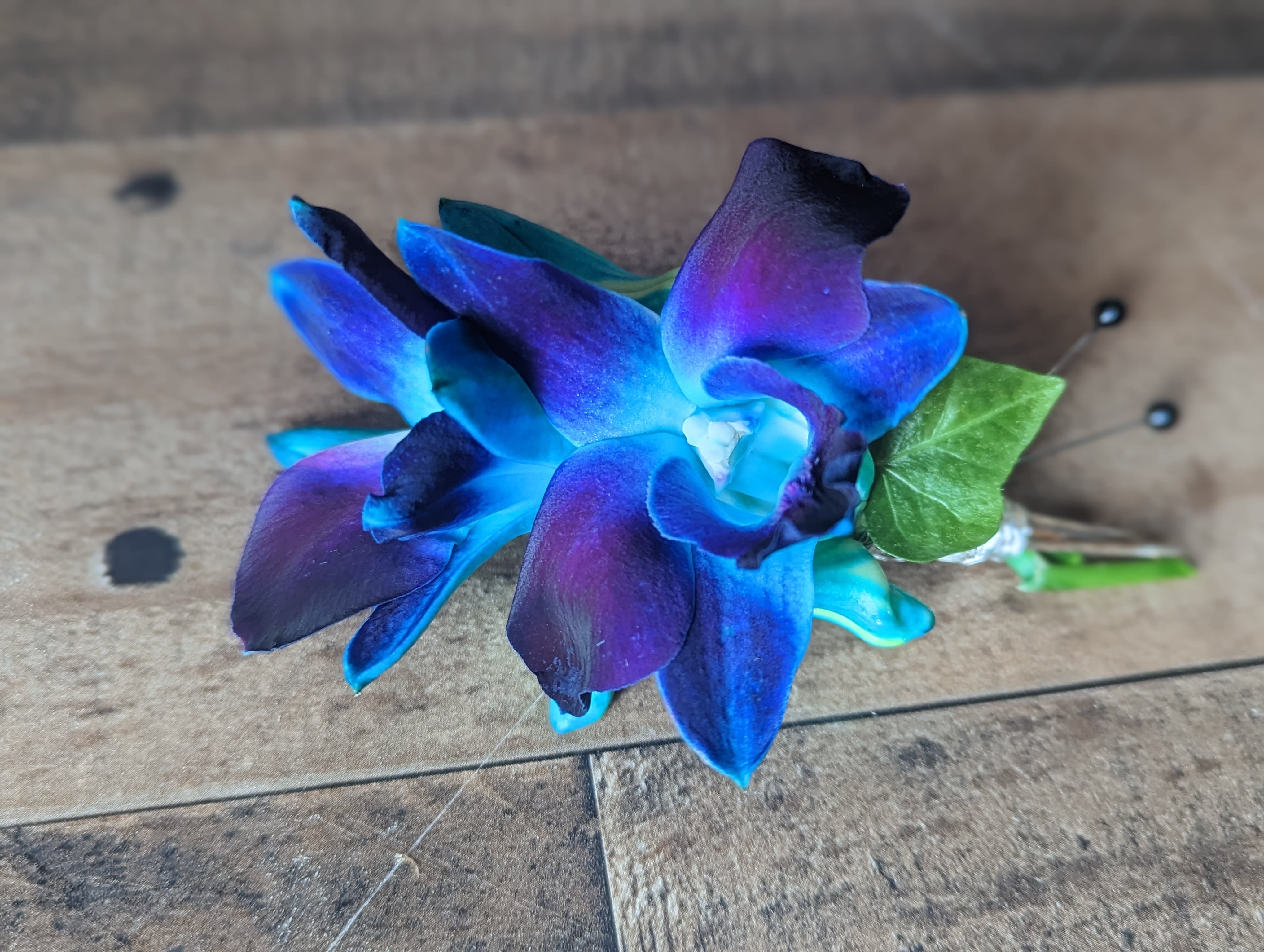 Blue Bomb Dendrobium Boutonniere - Shown as Standard Blue bomb dendrobium orchids come with accenting ivy and silver wire. *Pick up only* *We custom design this arrangement and use the freshest flowers available the day of delivery. The arrangement in this picture is an example of the size and style and may not feature the exact product shown.*