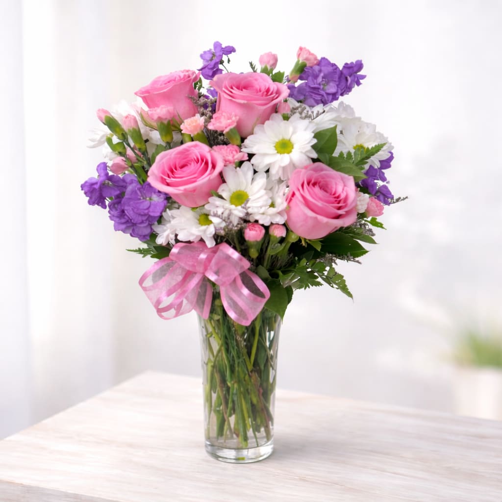 Blushing Romance - A joyful mix of classic red roses, crisp white daisies, soft pink mini carnations, and pops of playful purple stock — all gathered in a clear glass vase and finished with a flirty pink ribbon.