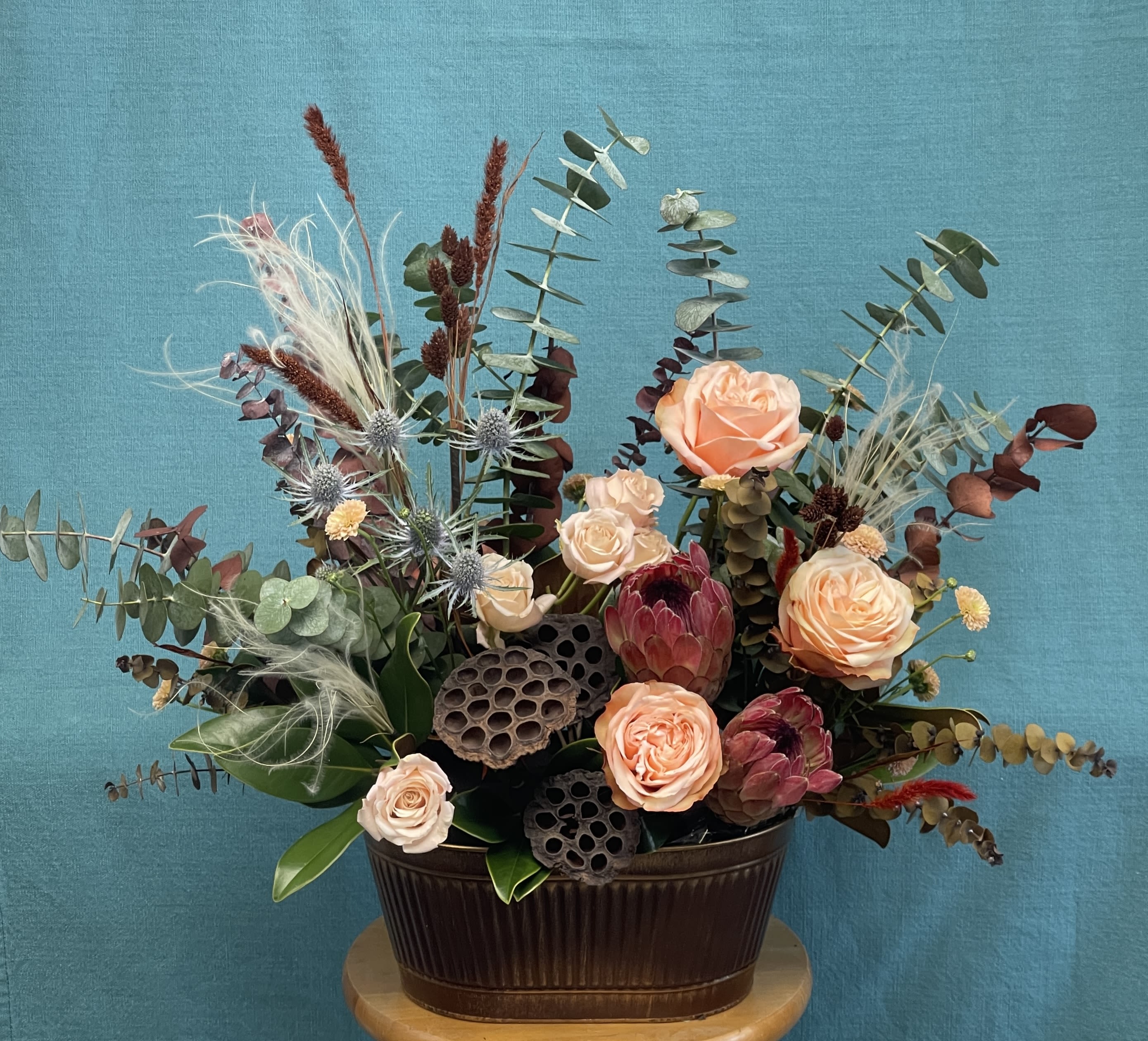Fall and Love - Autumn Elegance Arrangement Capture the warmth and texture of the season with our Autumn Elegance floral design. This rich arrangement combines soft peach roses, blush spray roses, and striking protea, beautifully paired with eucalyptus, lotus pods, and unique textural accents. The balance of warm tones and earthy elements creates a natural, sophisticated look perfect for any setting.  Designed in a deep ceramic container, this piece is ideal as a centerpiece for gatherings, a seasonal gift, or a stunning accent for your home décor.  Size: Approximately 20” wide x 18” tall Style: Lush, textural, and modern rustic 