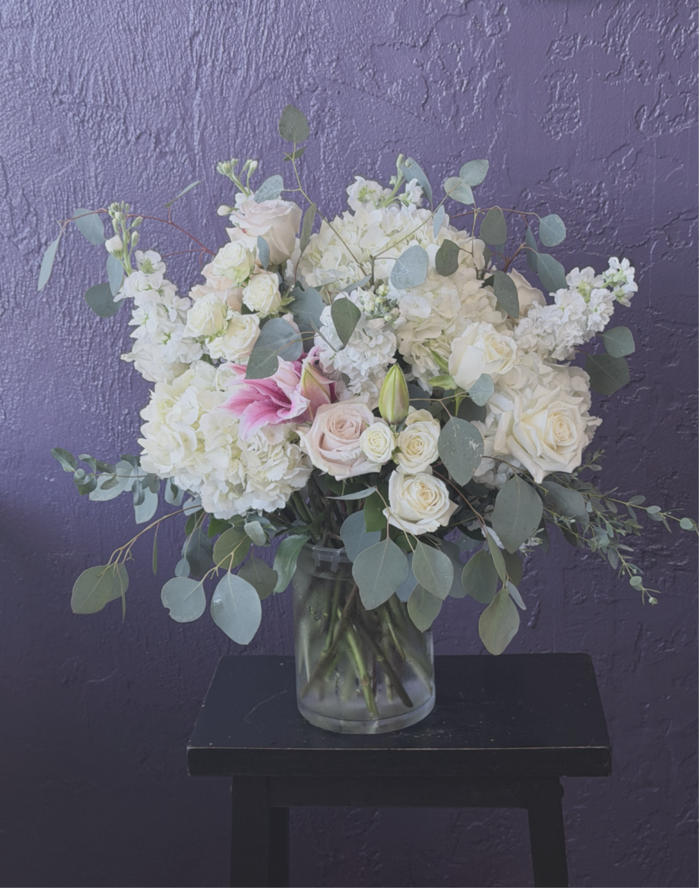 White and blush bouquet of roses, hydrangeas, and lilies in a clear vase against a purple wall