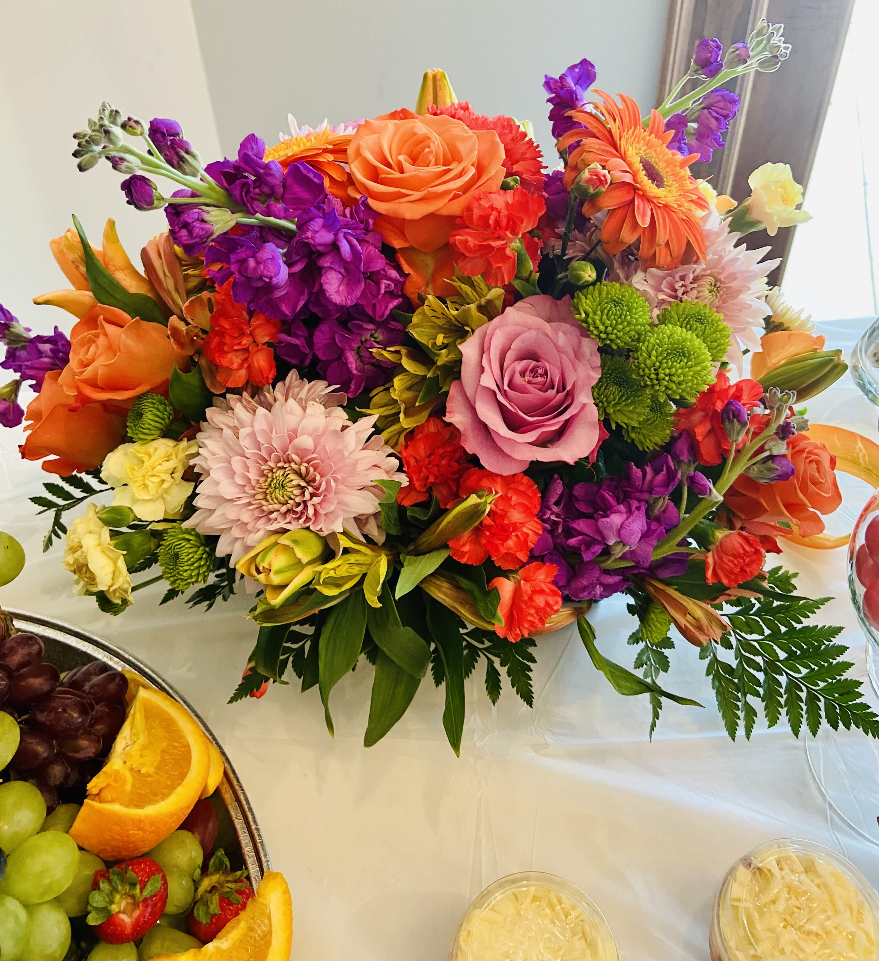 Just Because Basket by SWF - A lively, low-profile floral basket that makes someone feel noticed and celebrated-perfect as a just because arrangement, birthday bouquet, or a bright anniversary gift. Bursting with orange roses and lavender-pink roses, this tabletop display blends cheerful pink chrysanthemums, deep purple stock, and green button mums so the recipient feels genuinely seen the moment it arrives. The mix reads playful and upbeat, ideal for saying "I'm sorry," "congratulations," or "thinking of you" - and it also fits Mother's Day without feeling seasonal.  This compact design sits in a low container and is shown here styled with a serving tray of fresh fruit and glass bowls, making it a natural centerpiece for casual gatherings or a special breakfast surprise. Textural layers of orange gerbera daisies, vivid red-orange carnations, yellow alstroemeria, and clusters of stock create a full, rounded silhouette that looks abundant from every angle. Order for delivery to mark a birthday, Best Friends Day, Boss's Day, or to simply brighten someone's day-its approachable size and bold color mix suit tabletops and reception desks alike.