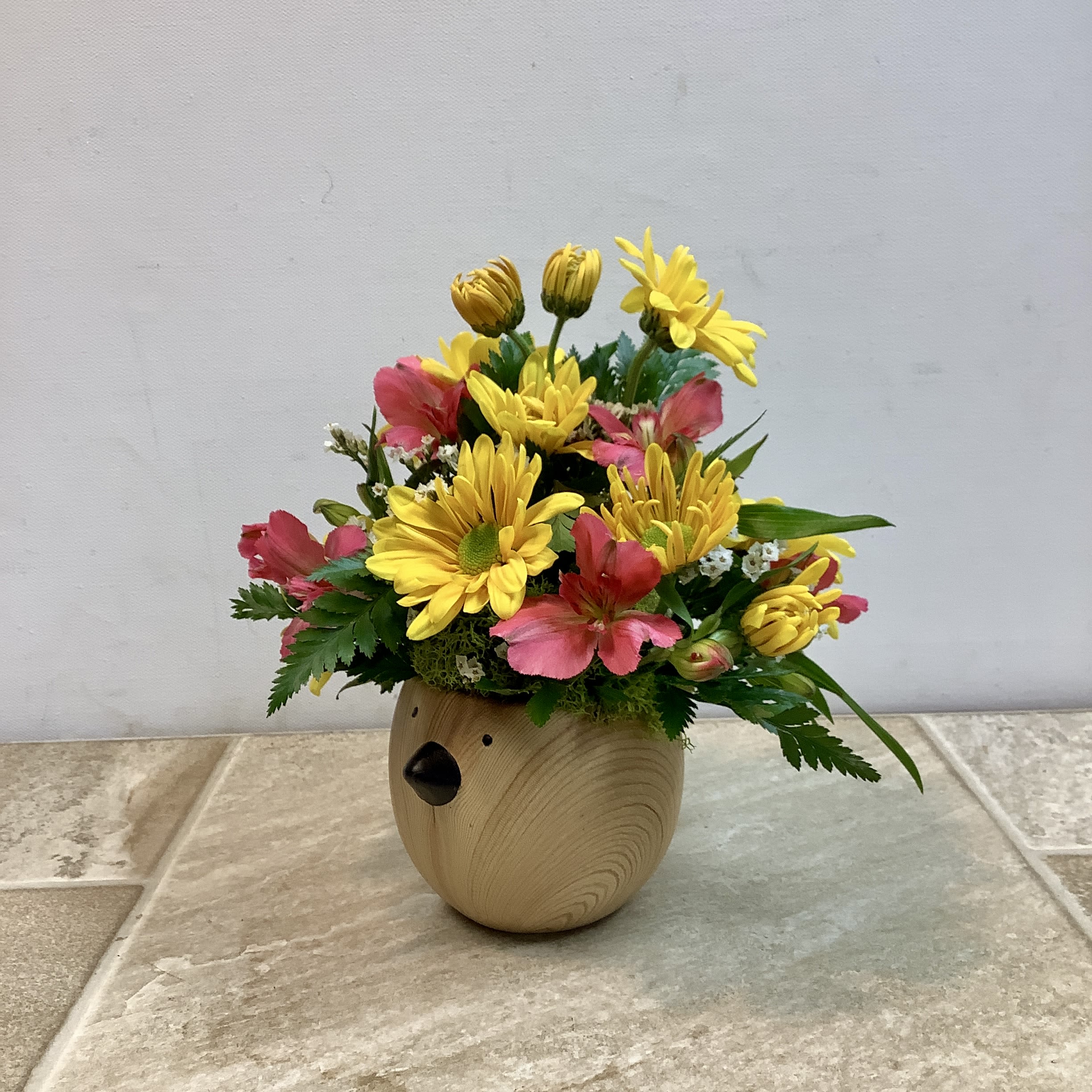 Chirpy Charm - This sweet little arrangement features sunny yellow daisy mums nestled among soft pink alstroemeria, delicate white filler flowers, and fresh greenery. The blooms are gathered compactly in a charming round ceramic bird container with a natural woodgrain finish and a tiny black beak, giving the piece a warm, woodland feel. Bright, cheerful, and full of personality, it’s a perfect petite gift to bring a smile. It measures 8” wide x 8” high. 