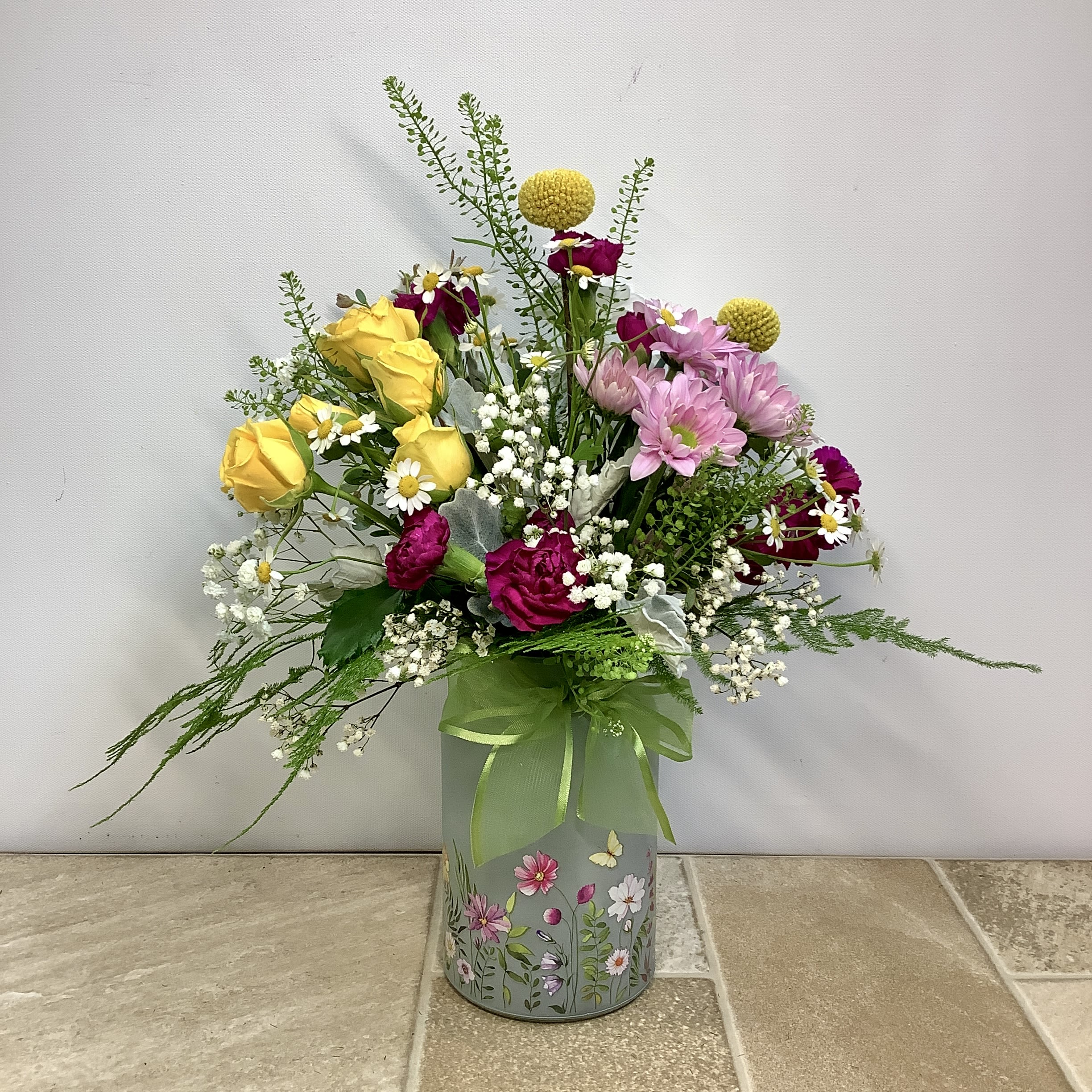 Marci’s Meadow  - Marci’s Meadow is a lovely, garden style bouquet with a joyful and colorful mix of blossoms that evokes the feel of a lively wildflower garden in full bloom.   The garden theme begins with a frosted cylindar vase with hand-painted-style floral decoration. It’s filled with a vibrant bright mix of cheerful yellow spray roses, white and lavender daisies, purple mini carnations, baby’s breath, and charming round Billy Balls (Craspedia). The natural, hand-picked look is enhanced with accent foliage like dusty miller, plumosa, pennycress, eucalyptus, and/or feverfew.       Approximate size: 13” wide x 16” tall.  Primary flowers: rose, daisy, carnation 