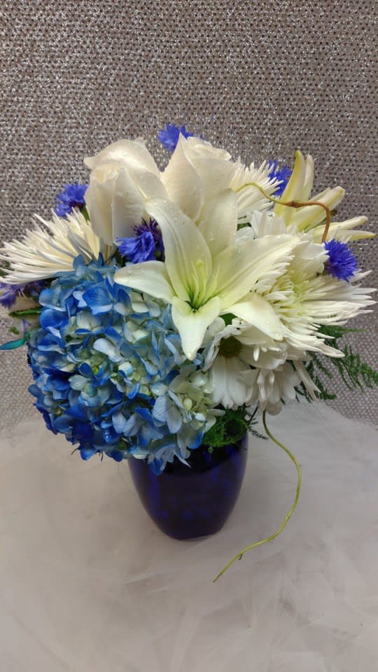 Bright Blues - This beautiful blue themed arrangement is made with white roses, hydrangeas, lilies, mums, daisy poms, and variegated filler greens. Dimension: 14"x10"