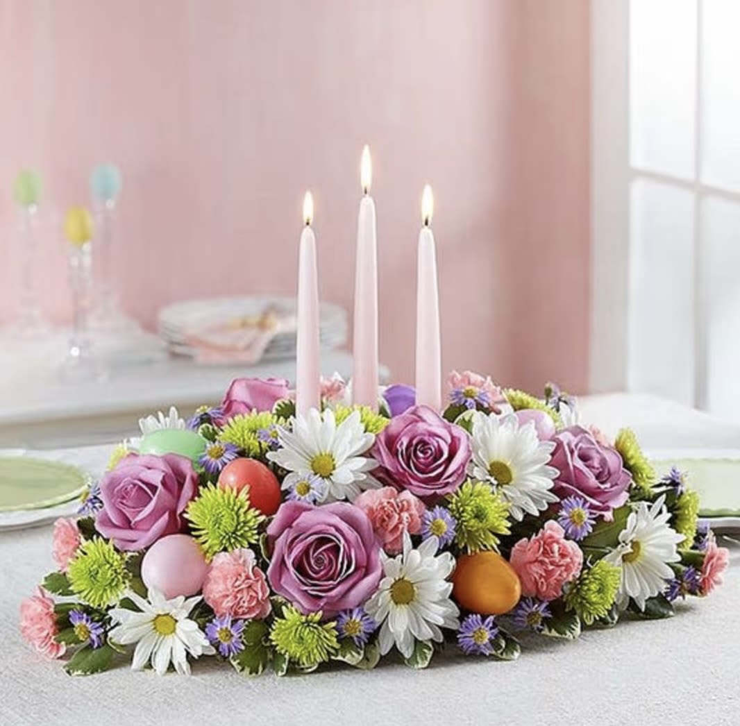 Easter Centrepiece  - At the heart of the table rests a low, elegant arrangement brimming with the beauty of spring. A mix of seasonal flowers spill gracefully across a natural base of lush greenery. The blooms are in a harmonious palette of soft pastels—blush pinks, buttery yellows, creamy whites, and pale lavenders—intertwined with vibrant pops of green foliage for a fresh, garden-picked look.  Nestled among the blooms are slender taper candles in soft ivory or pale blush, their gentle glow casting a warm, inviting light over the arrangement. The candles are carefully spaced to add ambiance without overpowering the flowers.