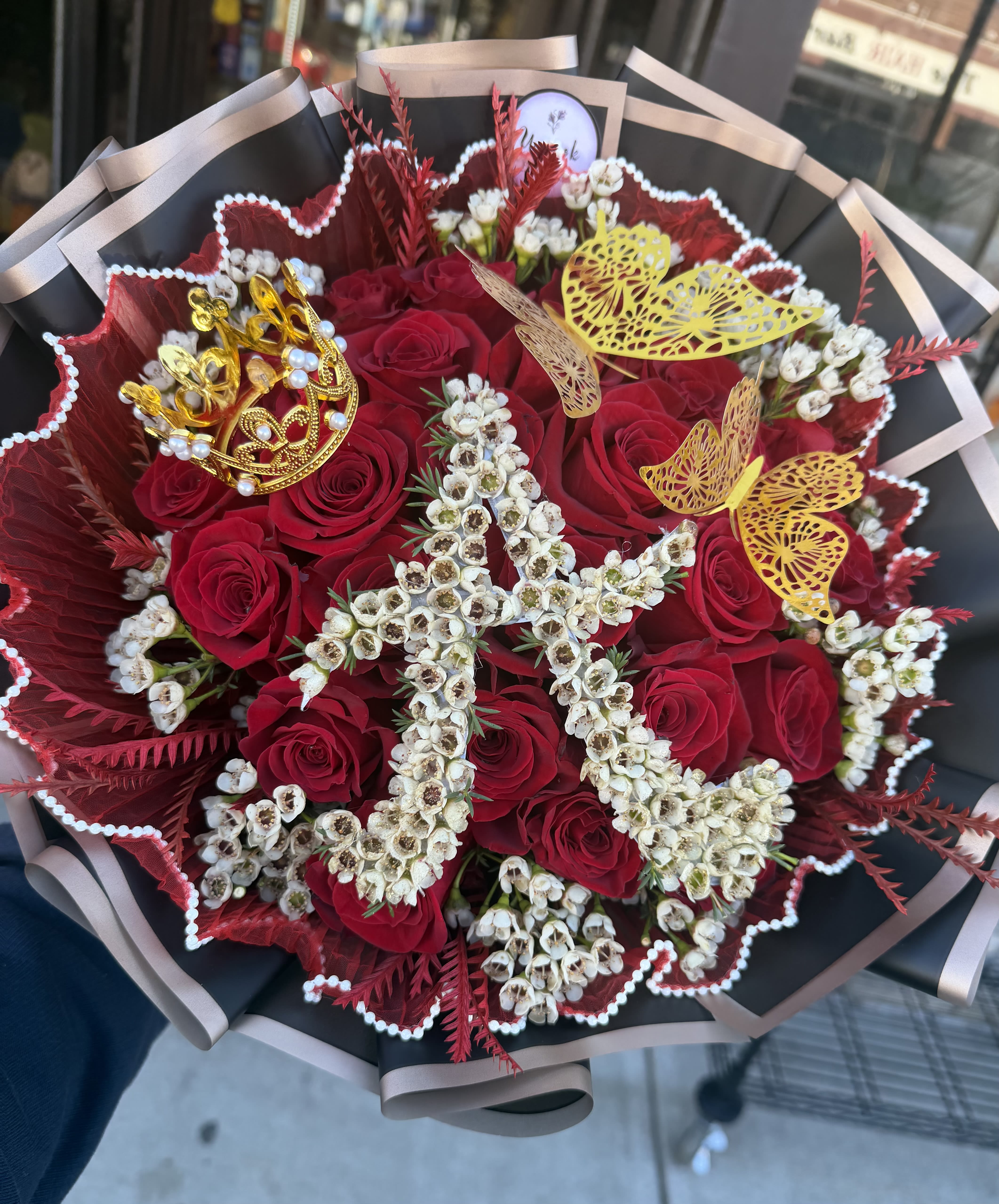 Hand-tied bouquet of red roses with white flowers shaped into a letter and gold crown and butterfly accents