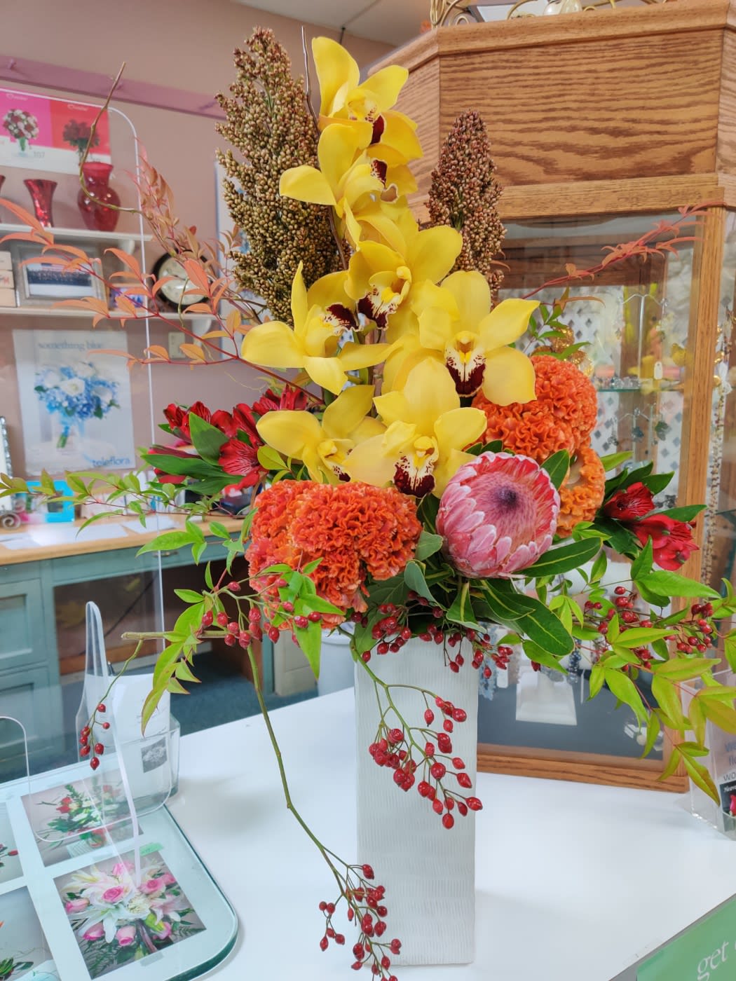 Tall arrangement with yellow orchids, orange blooms, pink protea and red berries in a white rectangular vase.
