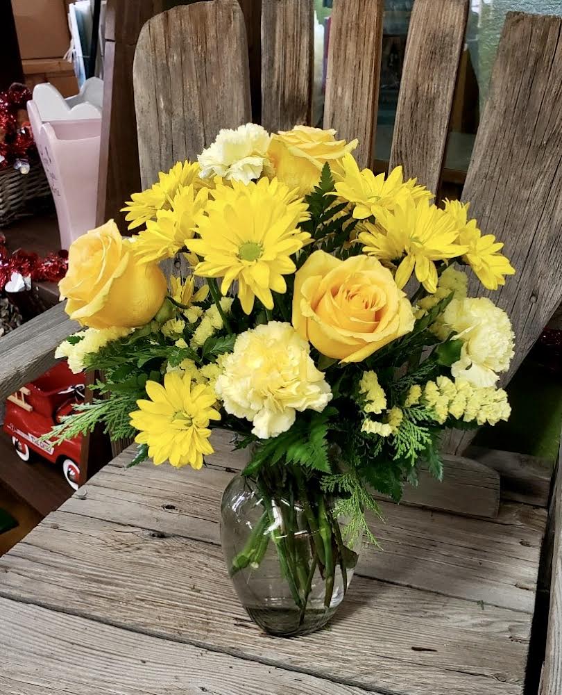 Yellow roses, daisies, and carnations arranged in a clear glass vase on a rustic wooden surface