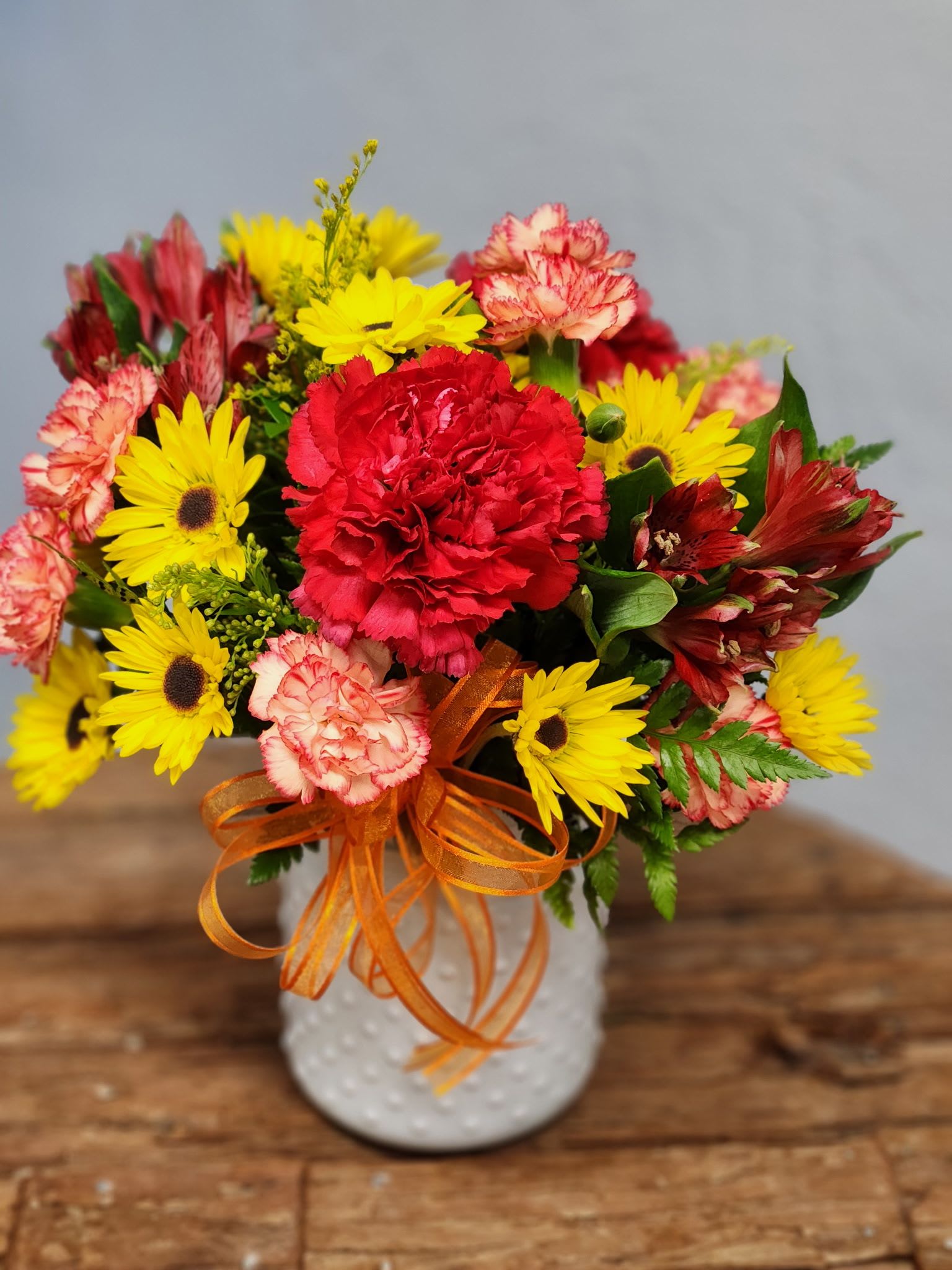 Bright arrangement of red and pink carnations with yellow daisies in a white vase with orange ribbon
