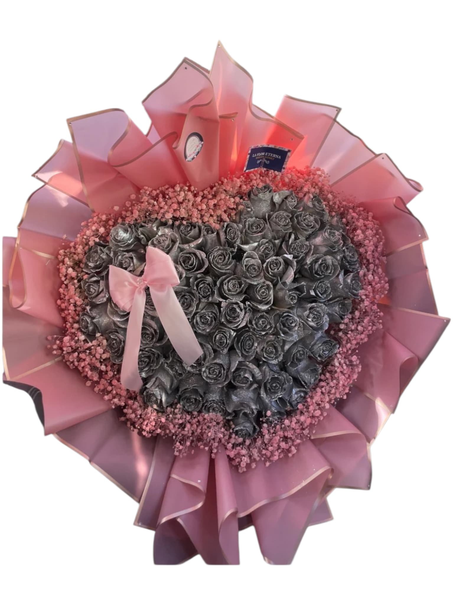 Silver Love - A dense heart of metallic silver roses steals the show in this heart-shaped flower arrangement, the perfect anniversary bouquet or Valentine's Day gift for someone who adores bold, modern romance. The cool, reflective petals create a dramatic focal point while a halo of tiny pink filler flowers softens the effect, so she'll feel both surprised and cherished the moment she sees it.  The blooms are wrapped in wide, ruffled pink decorative paper with a subtle edged detail that gives the arrangement a structured, present-like silhouette. A pale pink satin ribbon bow sits gently atop the roses, and small product tags finish the look for an easy, elegant presentation. Sized to be a clear statement piece, it arrives ready to display-no extra assembly required-making it an effortless way to say "I love you" with contemporary flair.