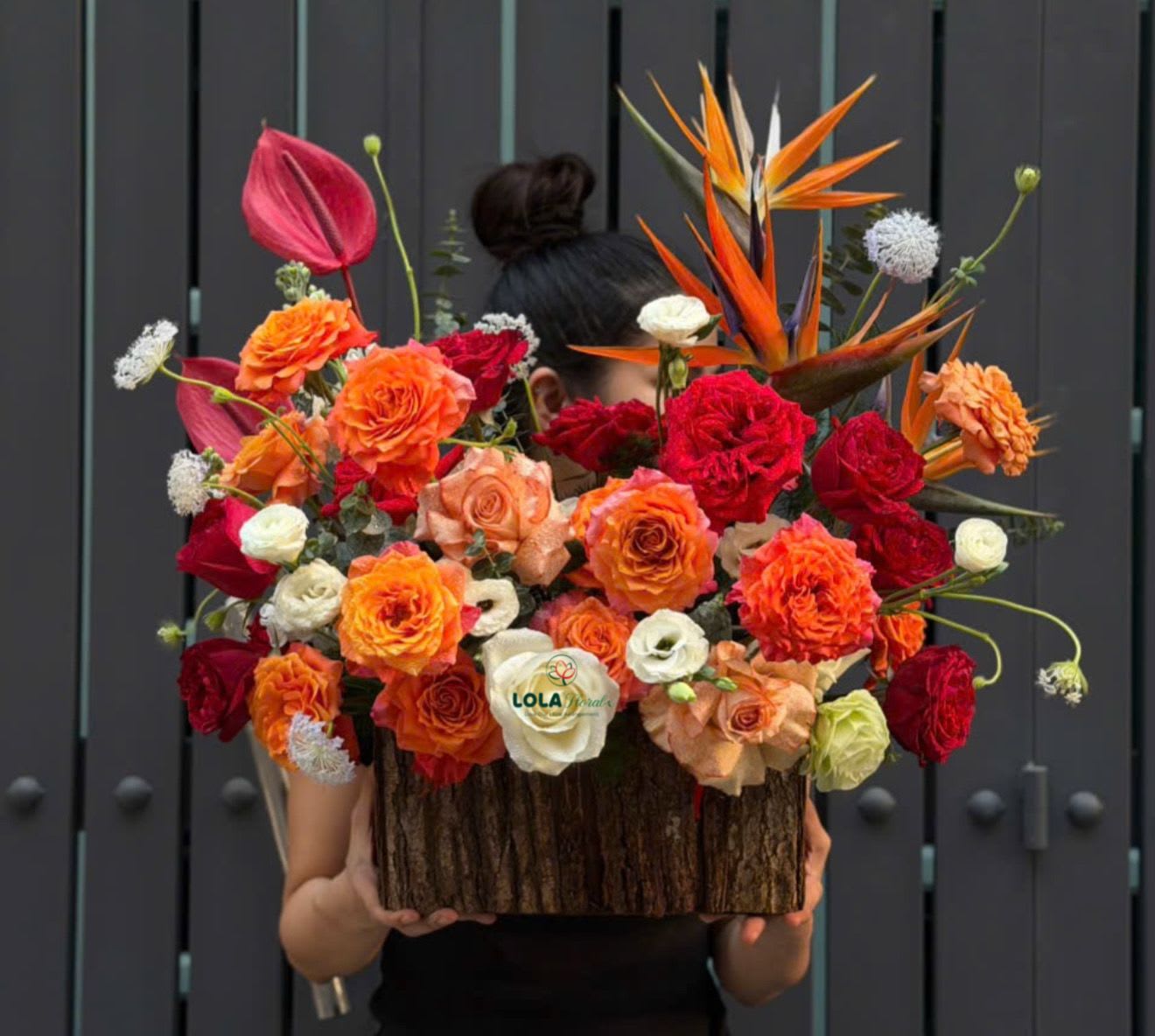 Fall Bloom - Vivid orange and deep red roses take center stage in this fall-inspired flower arrangement, a bold birthday or anniversary bouquet that feels celebratory and abundant. The rectangular wood container with a bark-like texture gives the design a rustic, seasonal grounding while tall bird of paradise stems and a pink anthurium add sculptural, tropical drama-perfect for someone who loves vibrant color and modern flair.  This lush display mixes roses, spray roses and tropical blooms in a palette of orange, red, peach and creamy white for striking contrast and depth. The dense massing of flowers creates a full, festive silhouette while the vertical lines from the bird of paradise and anthurium provide visual height. Arranged in a low, rectangular wood box, it reads both contemporary and cozy-an eye-catching centerpiece for a celebratory table or a thoughtful congratulatory gift.  Made to make the recipient feel truly celebrated, this arrangement suits birthdays, anniversaries, promotions, or any occasion that calls for warm, joyful color. The sturdy wood container arrives ready to display; keep blooms in fresh water and out of direct heat to extend their life.