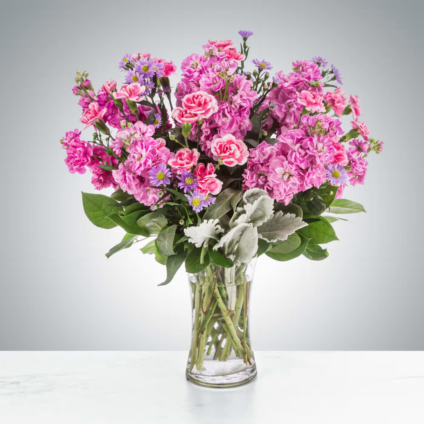 Tall bouquet of bright pink carnations, pink stock, and small purple asters in a clear glass vase.