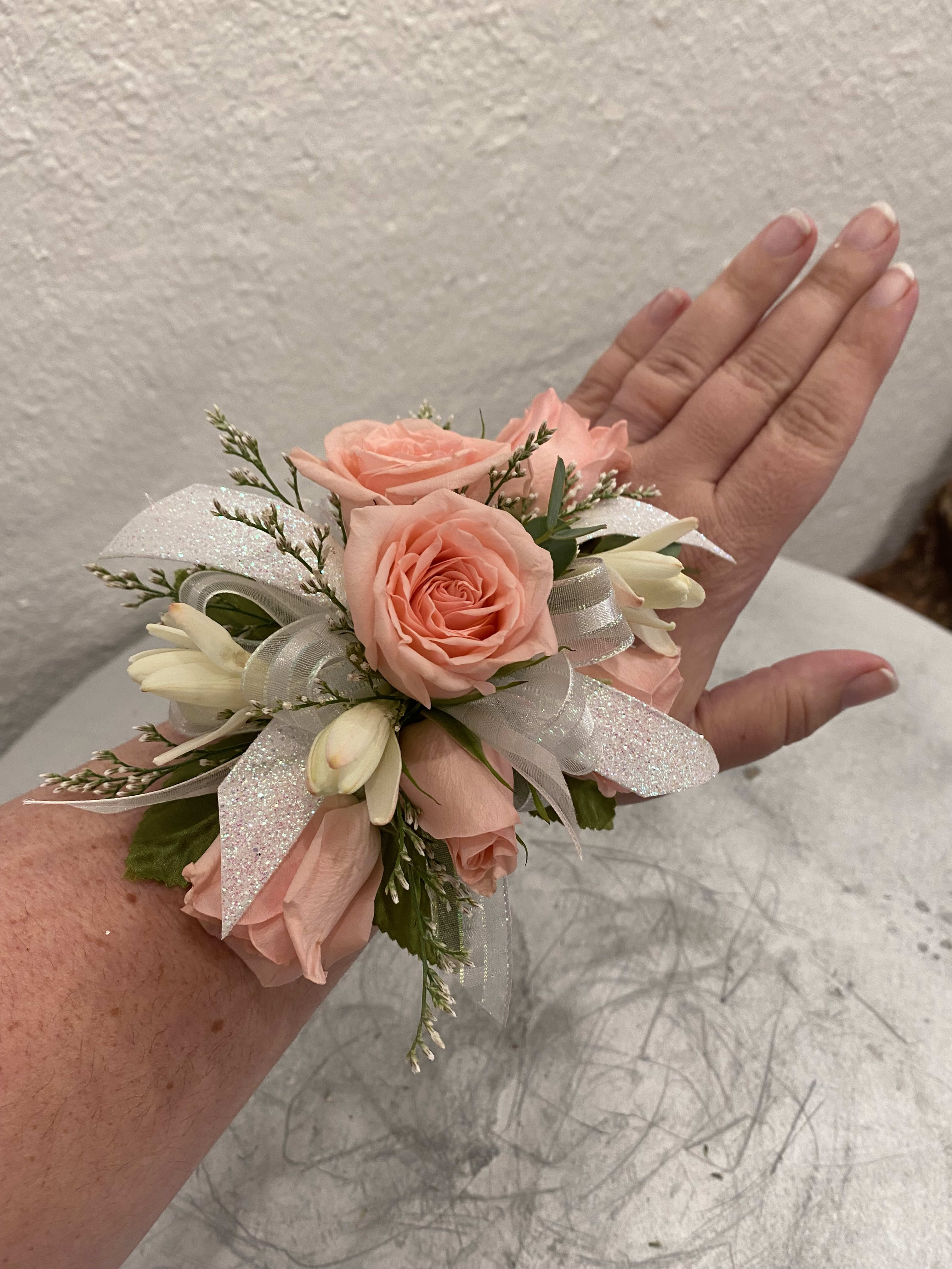Wrist corsage with peach roses, white buds, and sparkly white ribbon on an arm.