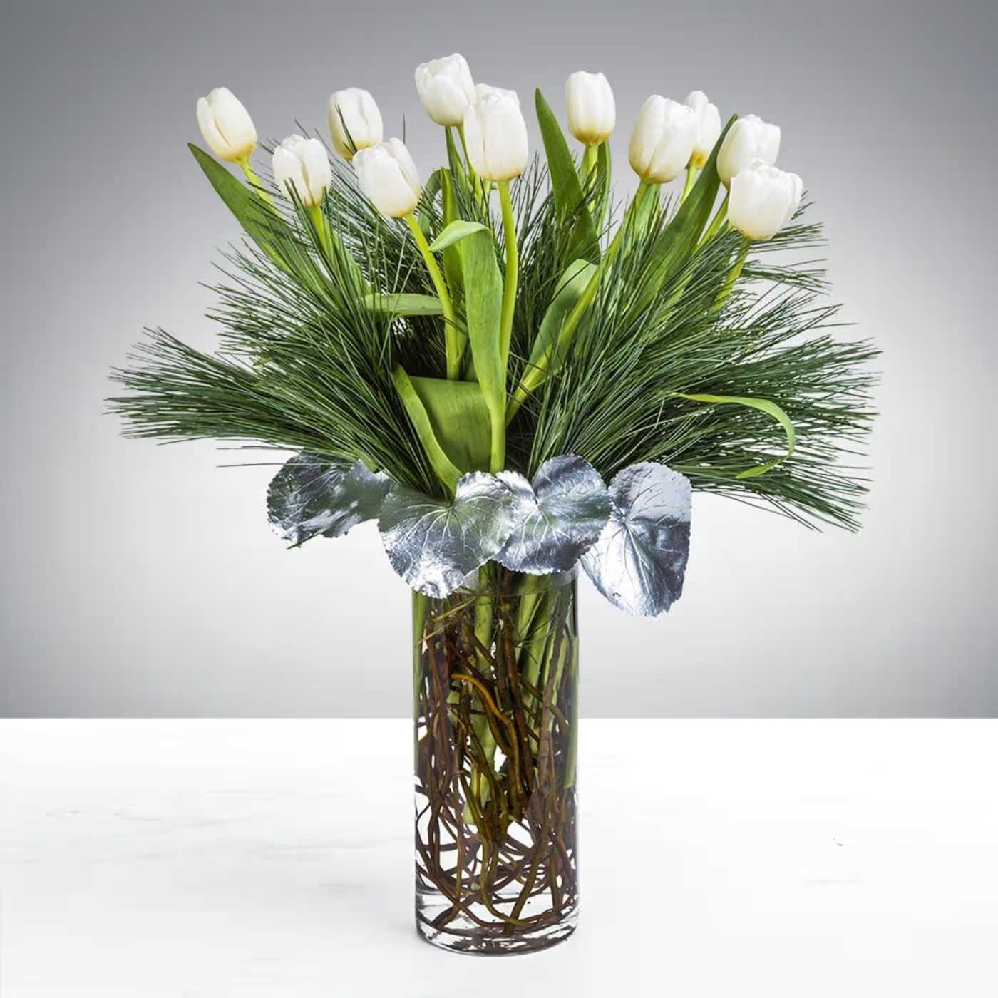 Tall clear vase with white tulips, pine needles, and silver accents in a modern arrangement