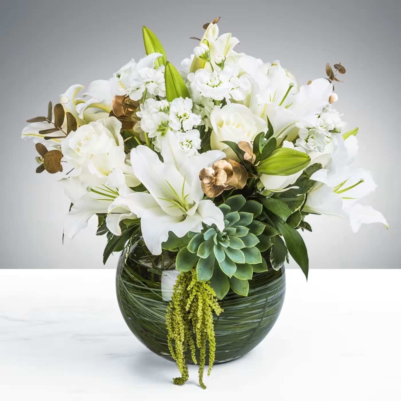White lilies and roses with succulents in a round green glass vase with trailing green accents