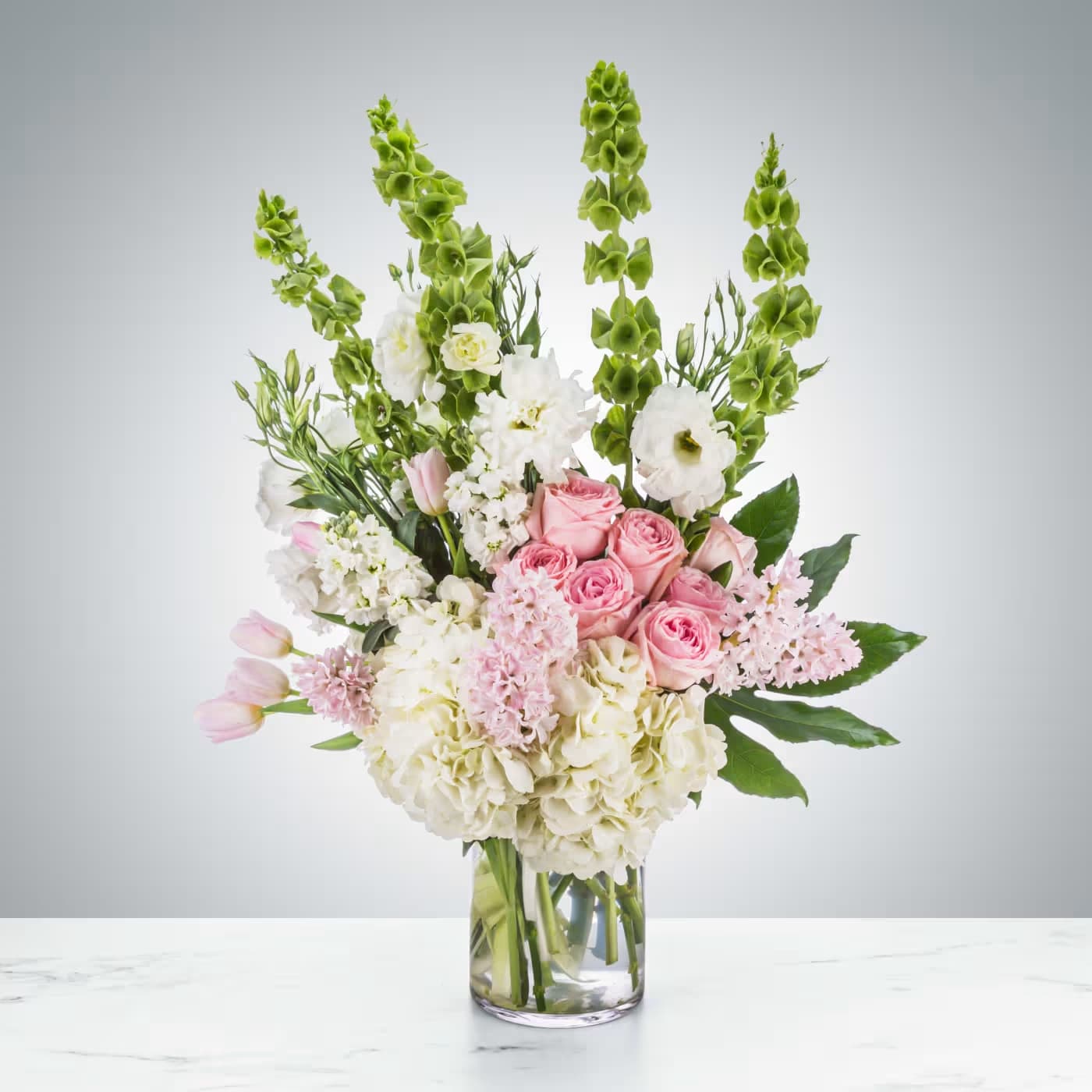 Tall arrangement of pink roses, tulips, and white hydrangeas with green bells of Ireland in a glass vase