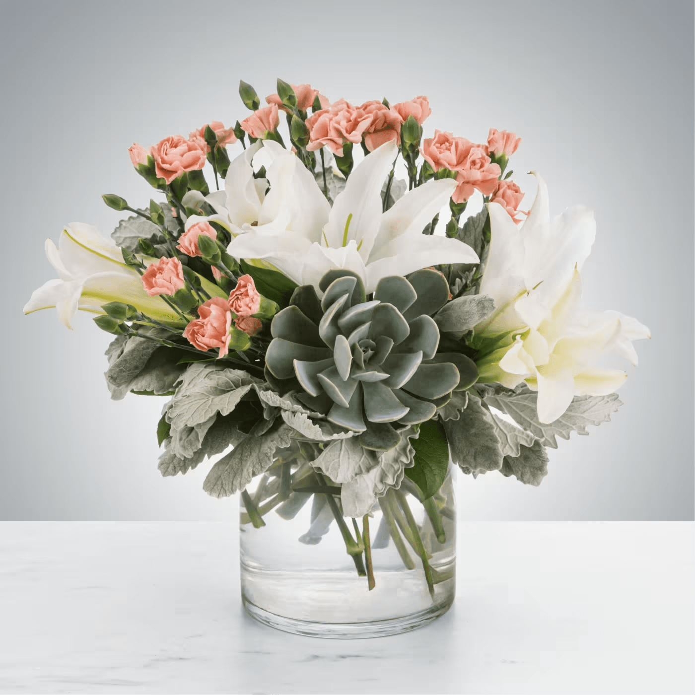 Arrangement of white lilies, peach carnations, and a central succulent in a clear glass vase