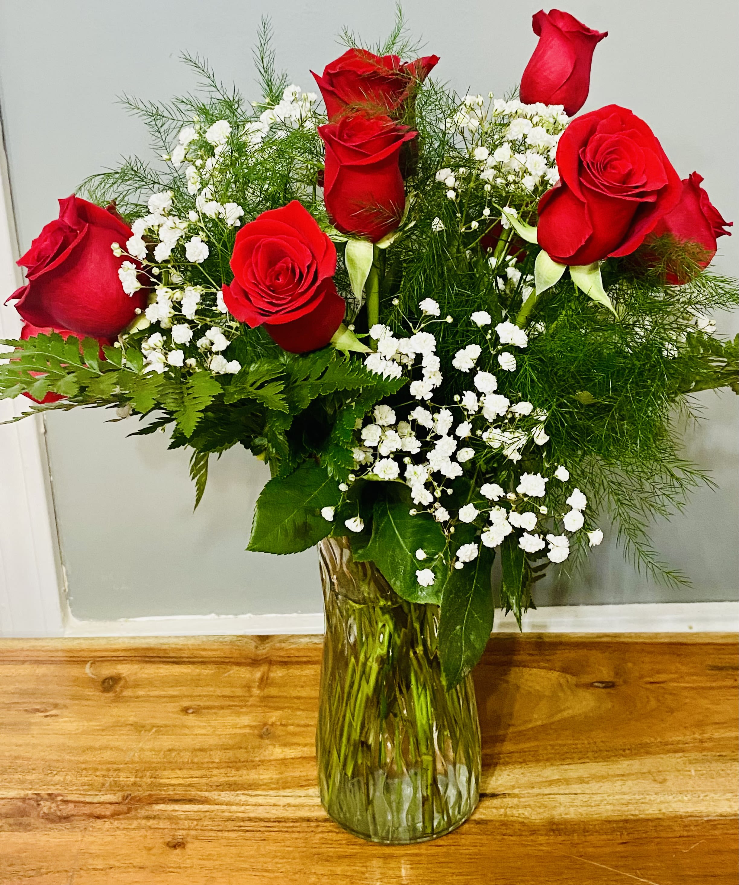 Classic Dozen Long Stemmed Red Roses in Vase - Long-stemmed red roses gathered in a clear glass vase make an unmistakable statement of love and romance-the kind of dozen roses that makes the person you're thinking of feel truly seen. This long-stemmed red rose arrangement is ideal for anniversaries, weddings, Valentine's Day, or simply a "just because" gesture that speaks from the heart.  Twelve classic red roses stand upright and radiate outward, accented with clusters of small white filler flowers that add delicate contrast and texture. Lush green foliage, including ferns and variegated leaves, creates a full, rounded silhouette that frames each bloom and enhances the roses' rich color. The bouquet is presented in a clear, textured glass vase for a timeless, elegant finish.  Perfect for saying "I love you," celebrating a milestone, or dressing a sweetheart's wedding table, this arrangement delivers classic romance with effortless style. Approximate dimensions: 25" H x 18" W; simple care-fresh water in the vase and a clean stem trim every few days will help prolong the life of the roses.