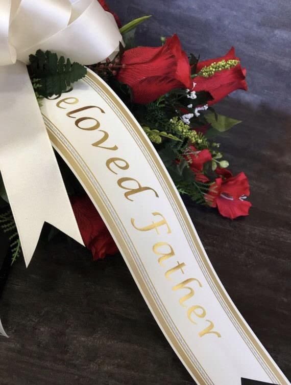 Sympathy spray with red flowers and a white ribbon reading Beloved Father, accented by a large ivory bow.