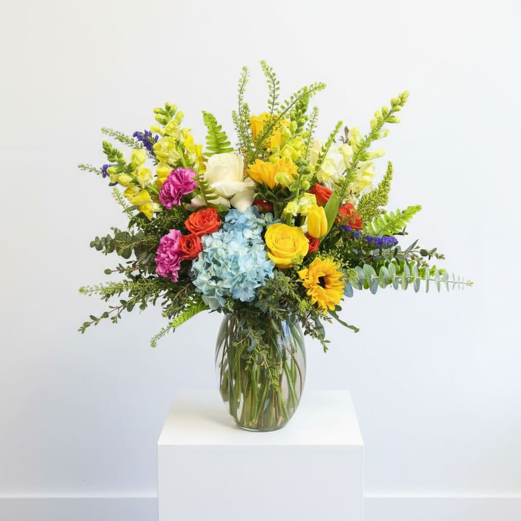 Sunshine And Grace - Bring joy and elegance into any space with the Sunshine &amp; Grace Bouquet—a radiant blend of hydrangeas, roses, sunflowers, tulips, snapdragons, ranunculus, stock, and lush greenery beautifully arranged in a glass vase. This stunning bouquet captures the essence of warmth, grace, and natural beauty, making it the perfect gift for any occasion.