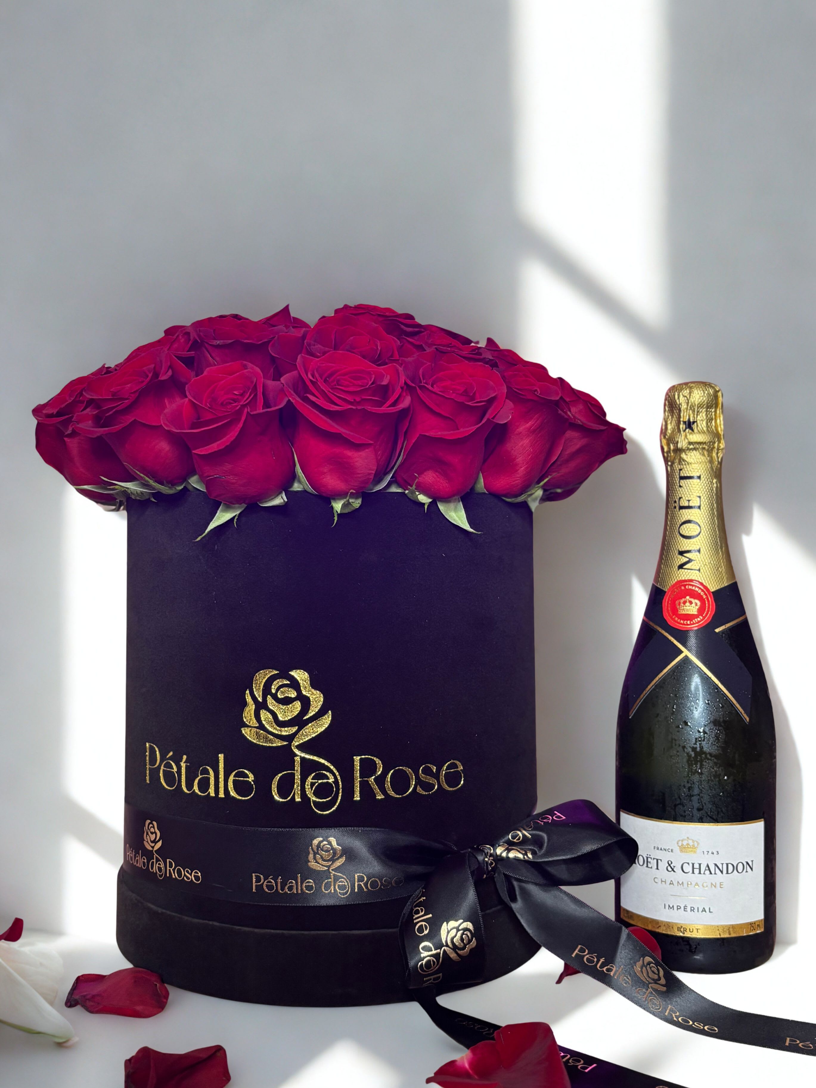 Black hat box of red roses beside a champagne bottle with scattered rose petals.