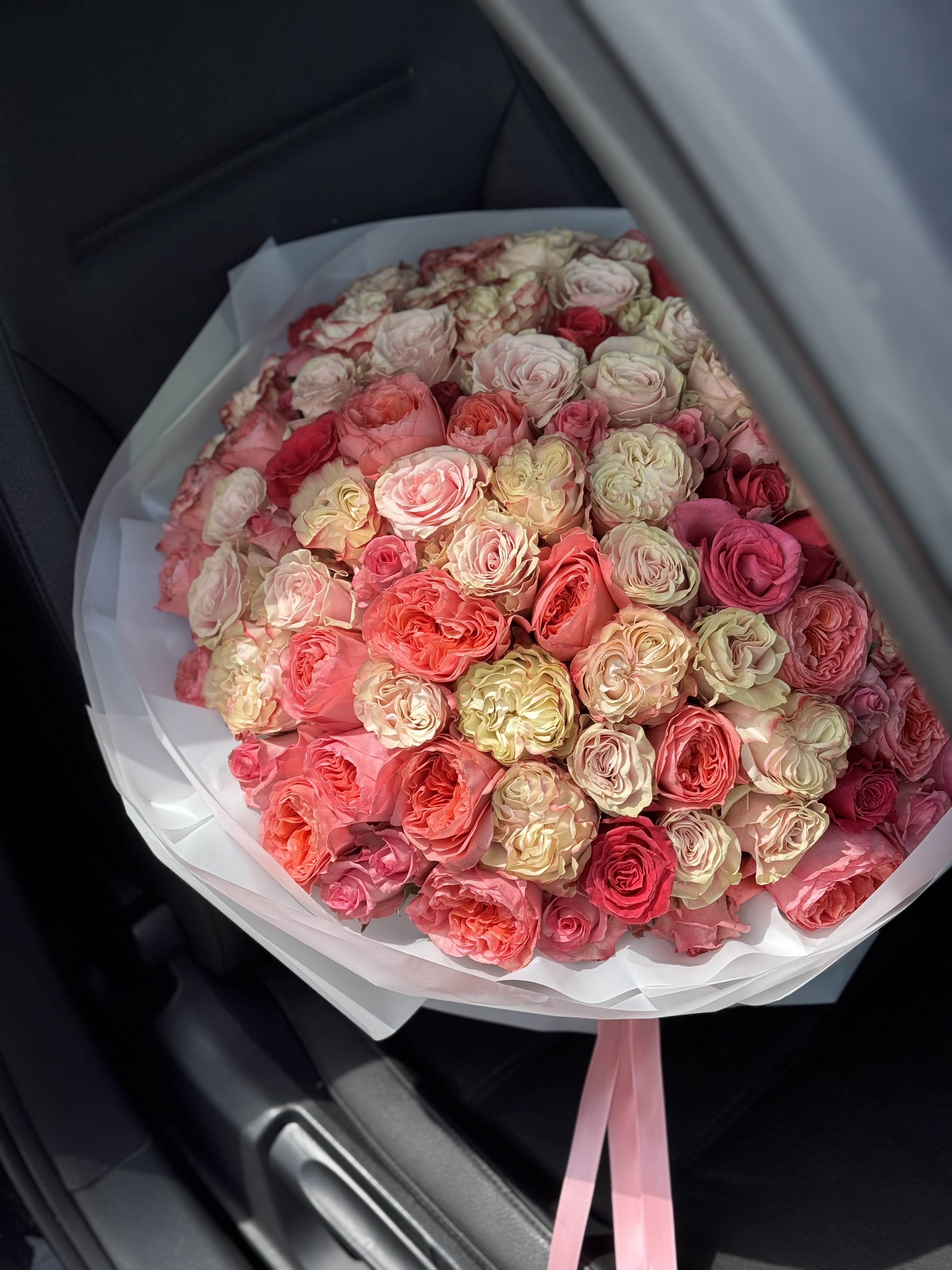 Large round bouquet of cream, peach, and pink roses wrapped in white paper with pink ribbons.