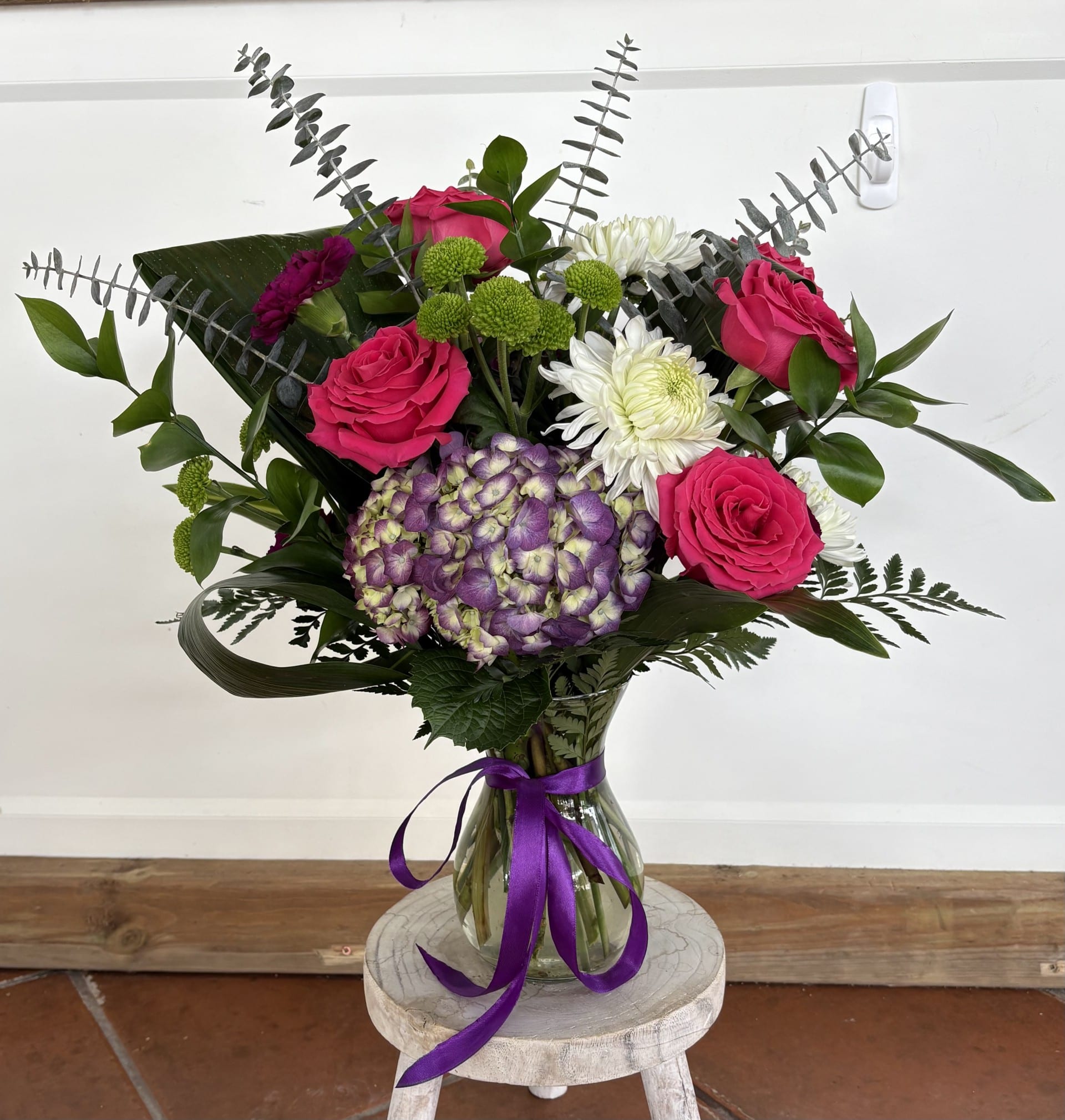 Lavender Love story - A lush, fan-shaped arrangement that reads like a Lavender Love Story-hot pink roses and a bold bicolor purple hydrangea take center stage, accented by white chrysanthemum-style blooms and small green button mums for playful contrast. This flower arrangement is perfect as a birthday bouquet, a congratulations gift, or a cheerful way to say you're thinking of someone special.  Tall silvery foliage and broad folded leaves give the bouquet structure and a sculpted silhouette, while the clear glass vase tied with a bright purple satin ribbon keeps the look fresh and polished. Shown here on a rustic wooden stool, the mix of textures-from velvety roses to fluffy hydrangea heads-creates a rich, tactile experience in pink, purple, white, and green.  Send this to mark milestone moments or to brighten someone's day; she'll feel celebrated and seen when these colors arrive. The arrangement comes gathered in the included clear vase with ribbon, ready to display-simple care: keep flowers in fresh water and out of direct sun to prolong bloom life.