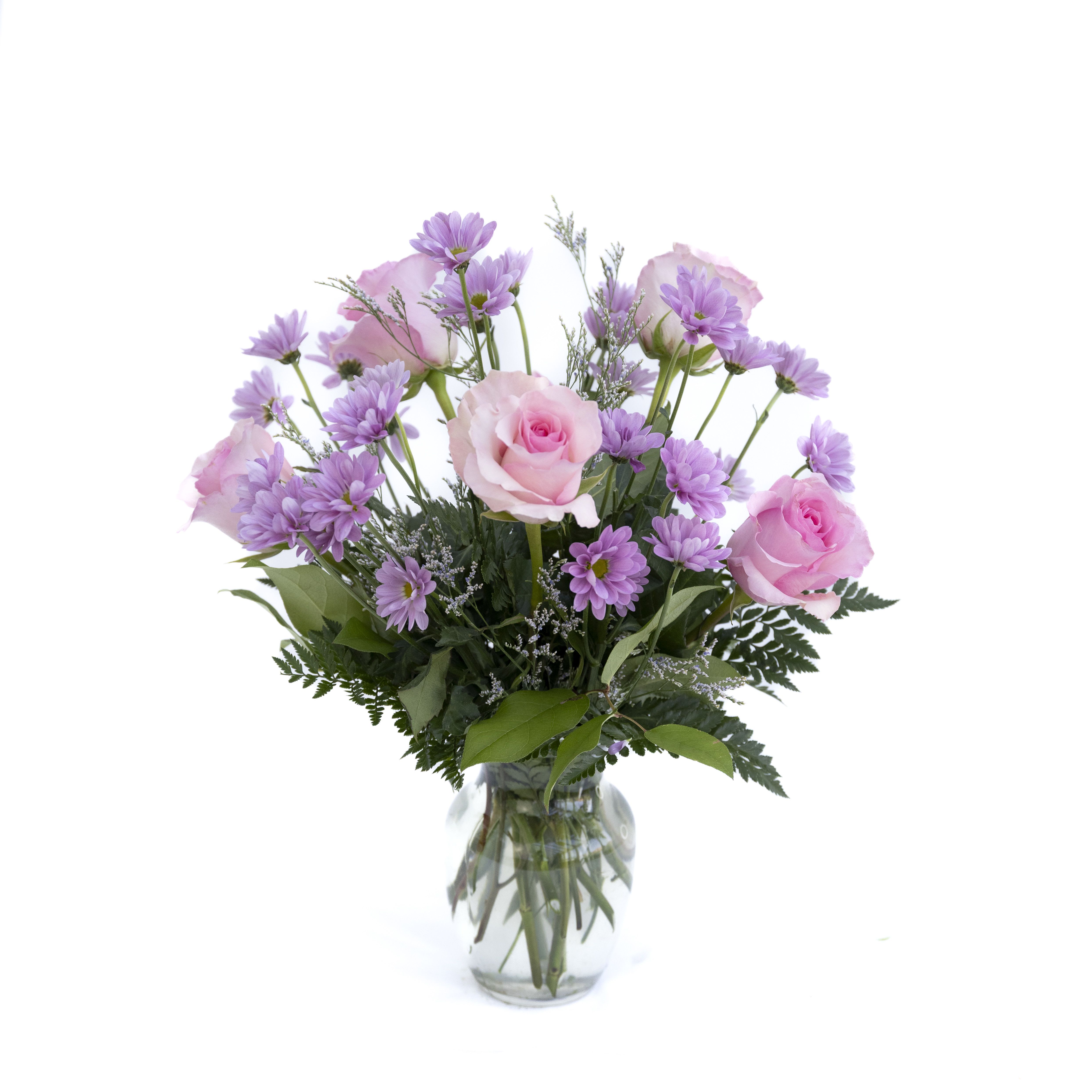 Blushing Blooms - Blushing Bloom is a sweet and charming arrangement designed in soft shades of pink and lavender. Featuring delicate pink roses paired with cheerful lavender daisies and airy accent blooms, this bouquet creates a light, garden-fresh feel. Arranged in a classic clear glass vase with lush greenery, it’s the perfect choice for celebrating love, friendship, or simply adding a gentle touch of beauty to any space. Soft, graceful, and full of charm, this arrangement is sure to brighten any day. 
