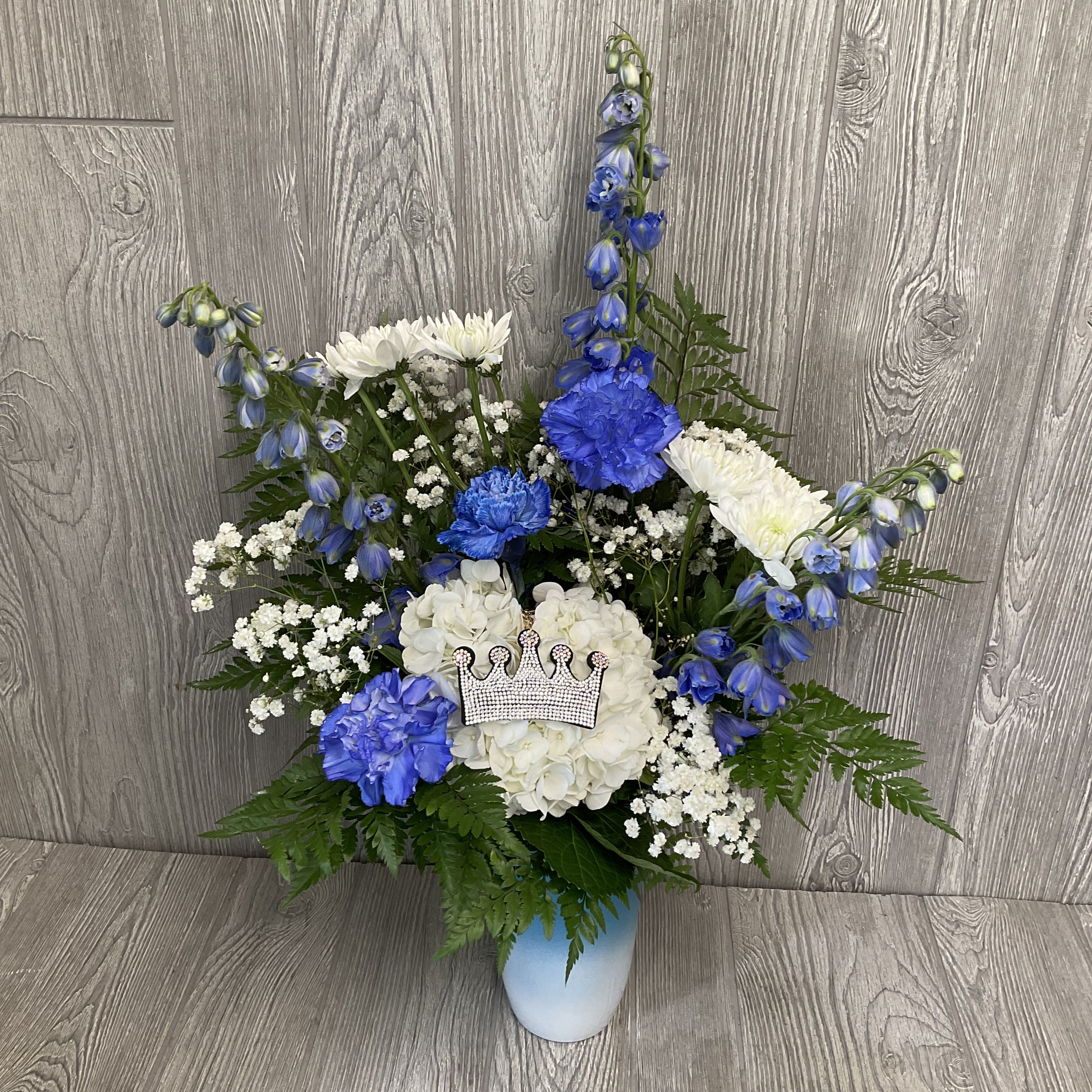 Tall blue and white flower arrangement with carnations, mums, hydrangea and a crown accent in a blue vase.