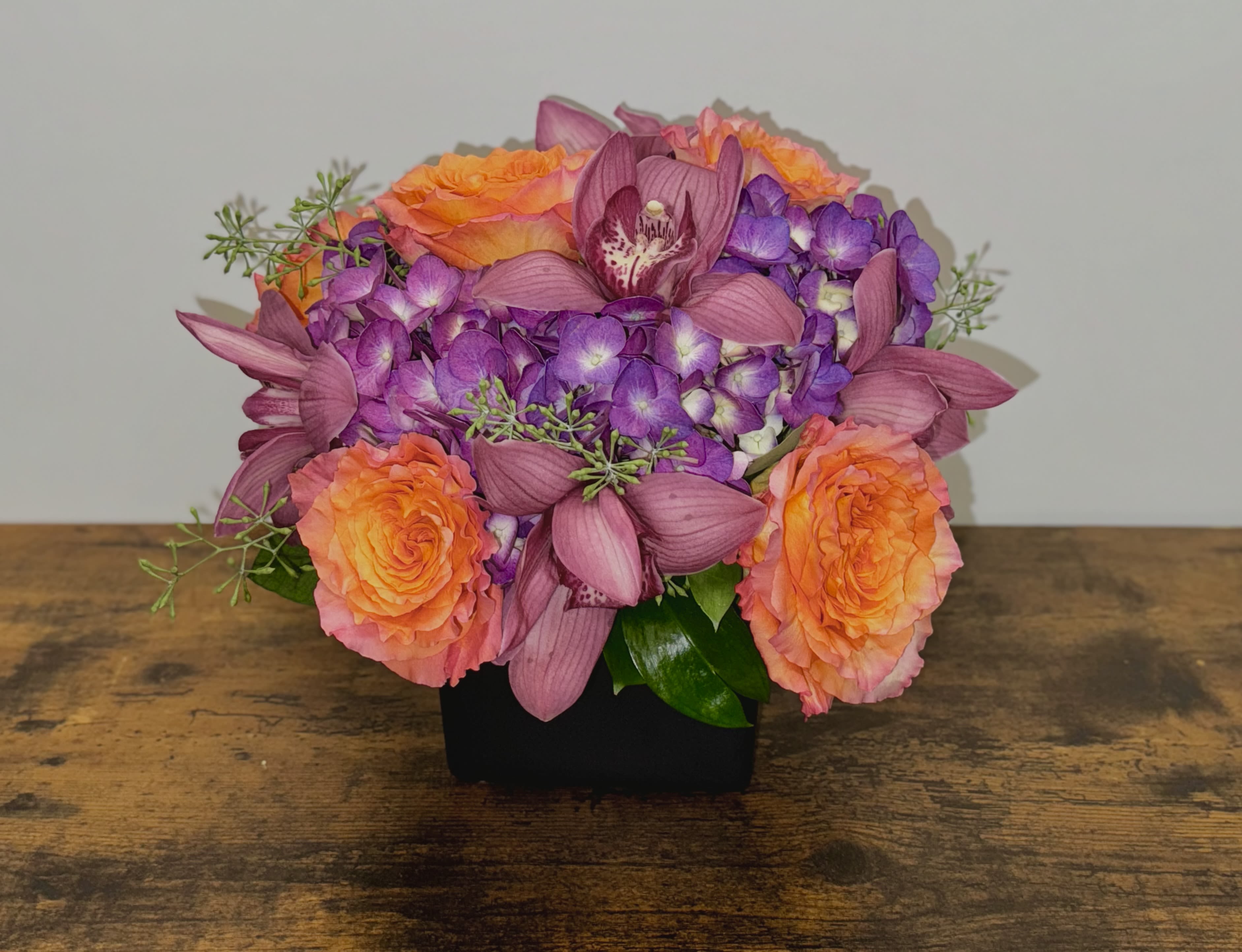 Low arrangement of orange roses, purple hydrangeas, and mauve orchids in a black square vase