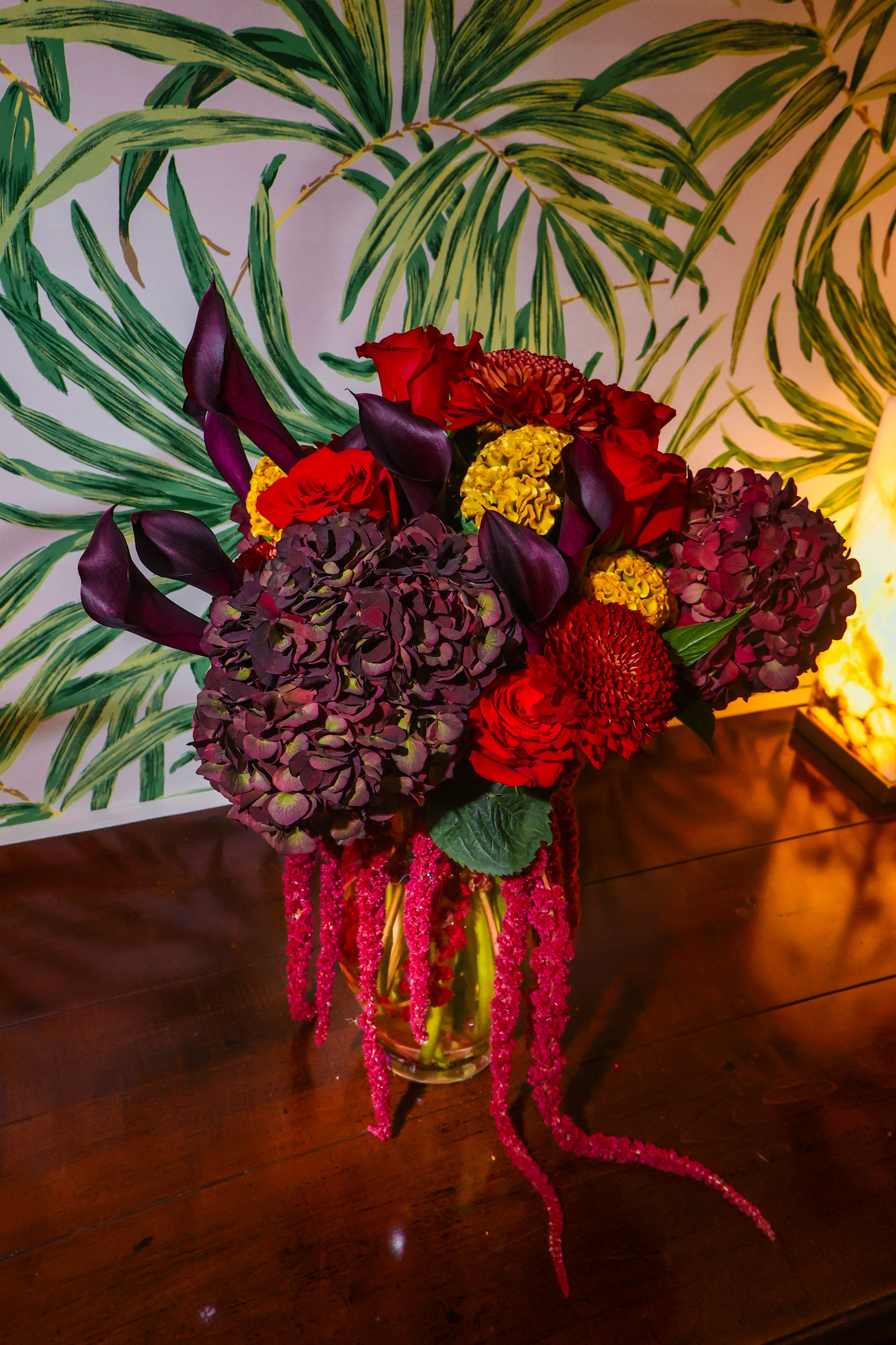 Jewel-toned arrangement of red roses, dark calla lilies, and hydrangeas with trailing pink accents in a glass vase.