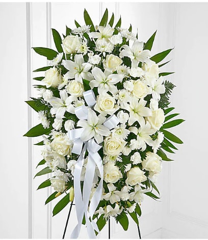 Tall standing spray of white lilies, roses, and carnations with white ribbon on a green leaf backdrop