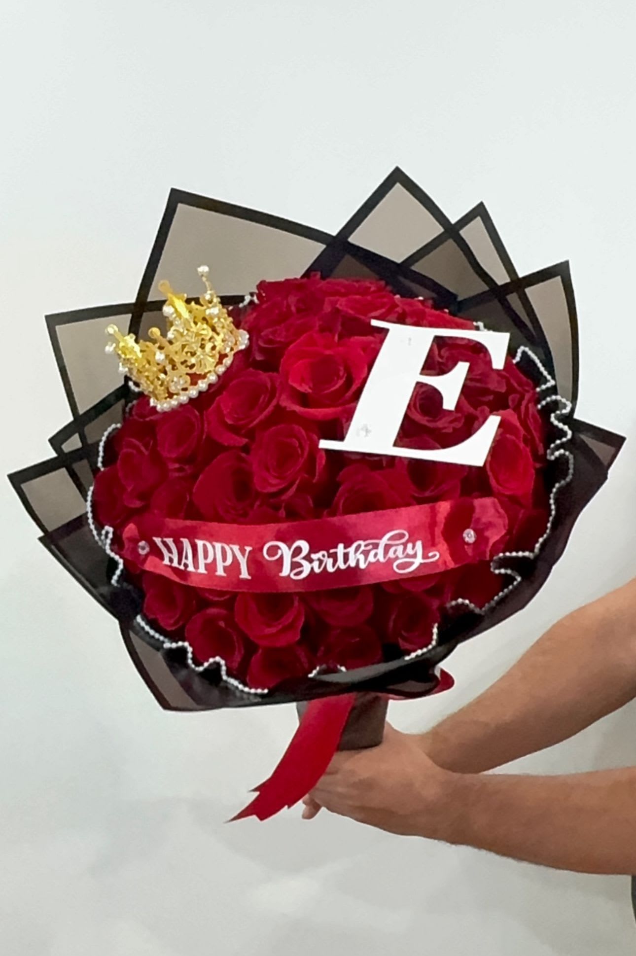 Bouquet of red roses wrapped in black paper with a crown, letter E, and Happy Birthday ribbon.