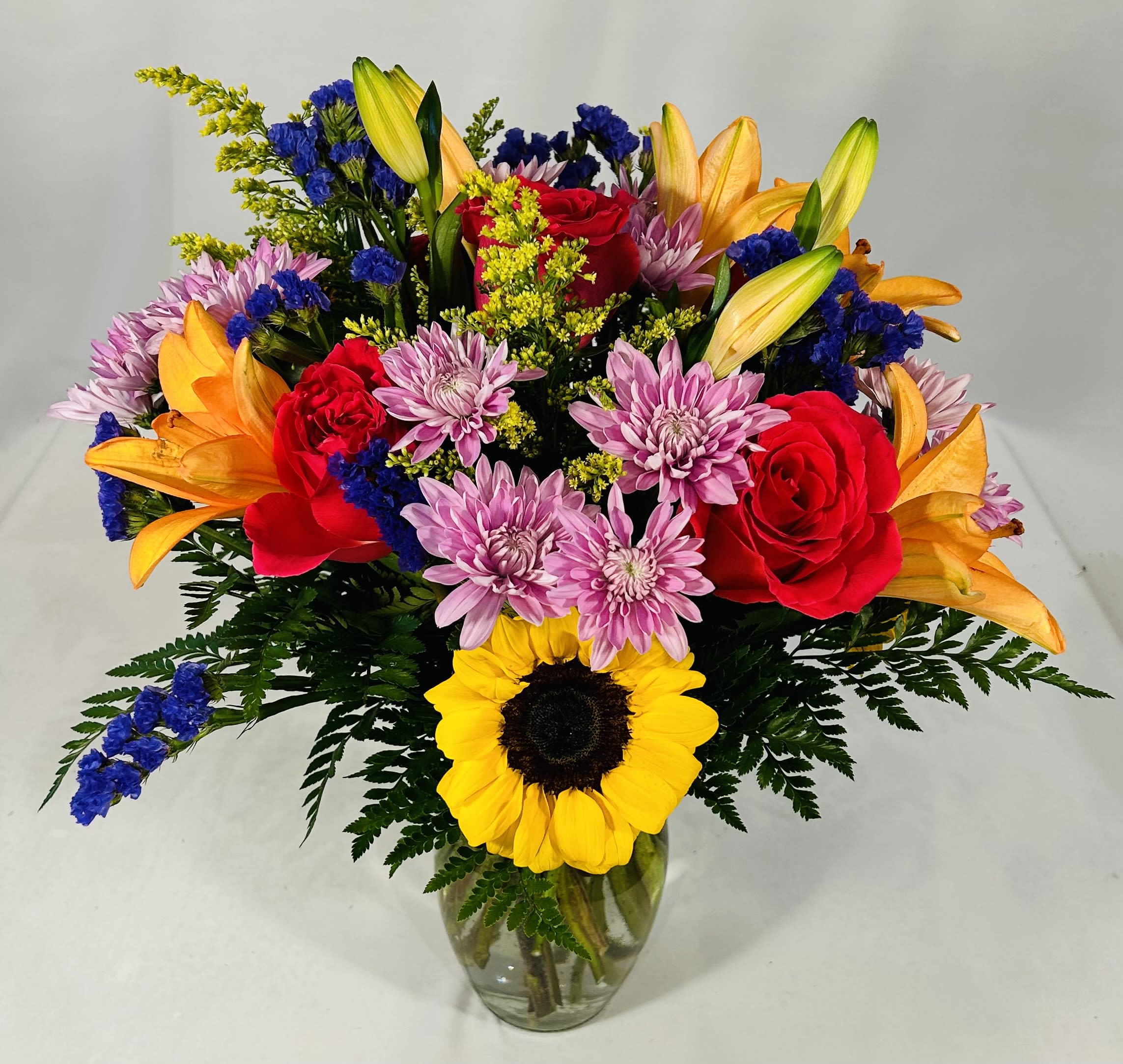 Bright mixed bouquet with sunflower, red roses, orange lilies, and purple mums in a clear glass vase