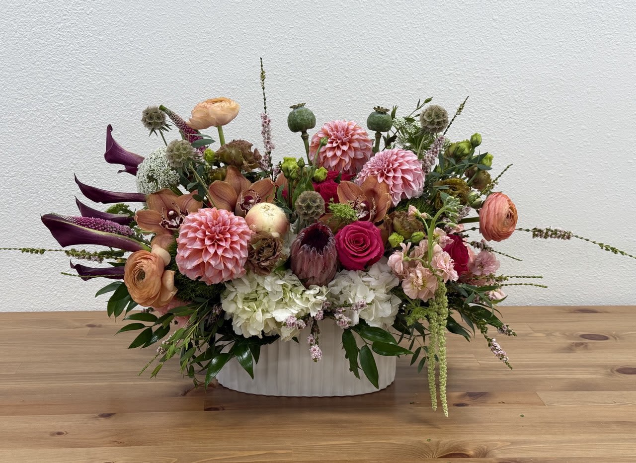 Low oval arrangement of pink dahlias, roses, calla lilies and mixed blooms in a white ceramic vase.