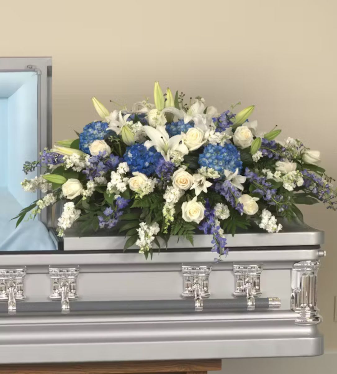 Large white and blue casket spray with roses, lilies, and hydrangeas on a silver casket.