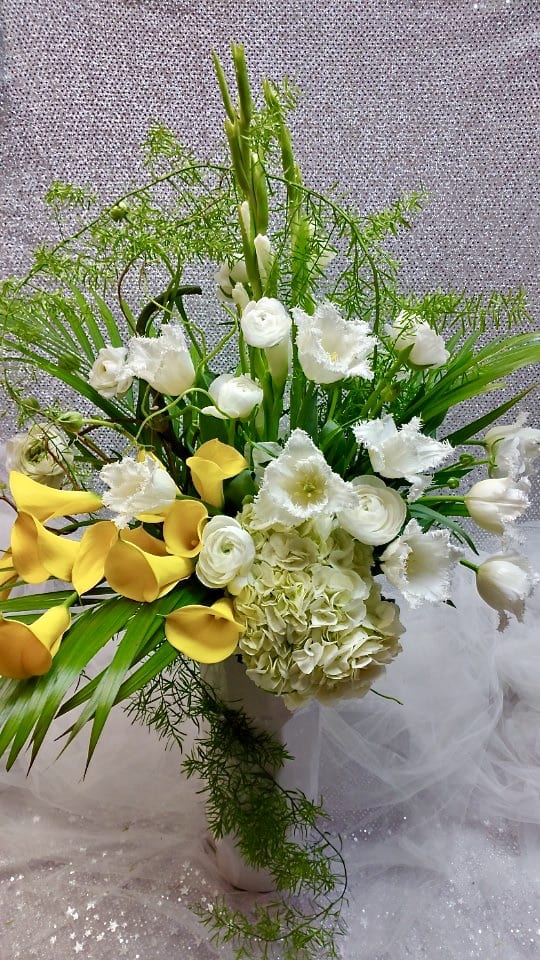 Tall arrangement of yellow calla lilies, white hydrangeas, tulips, and ranunculus in a white vase