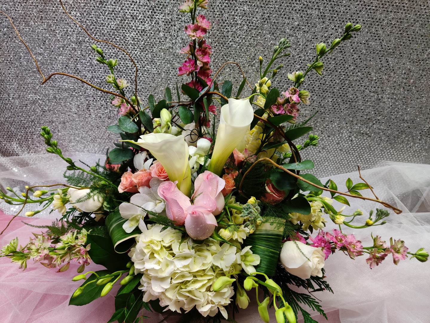 Low centerpiece of white calla lilies, hydrangeas, and pink roses with branches on a soft pink and silver backdrop