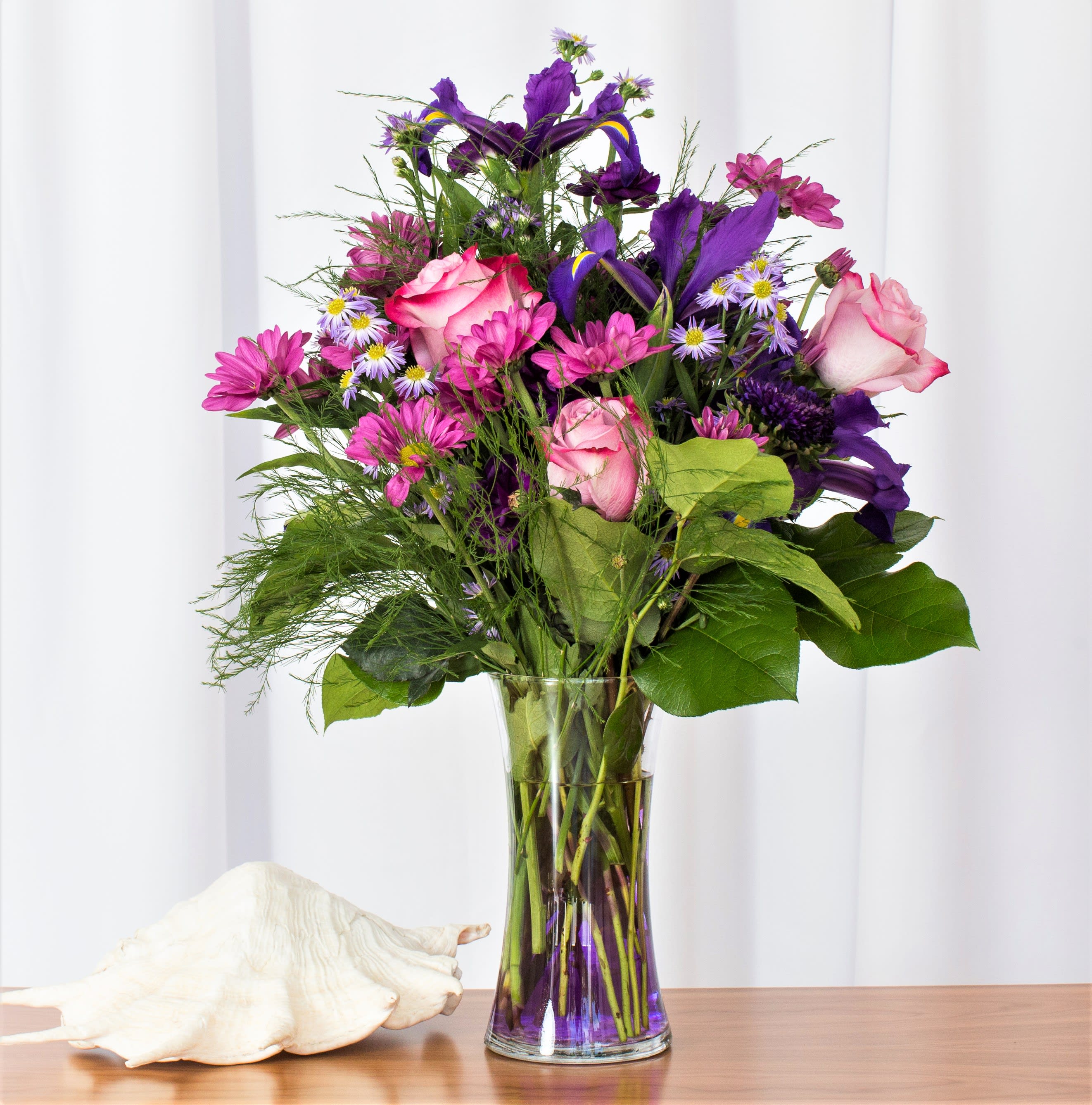 Tall bouquet of pink roses, purple irises, and pink daisies in a clear vase beside a large white seashell.