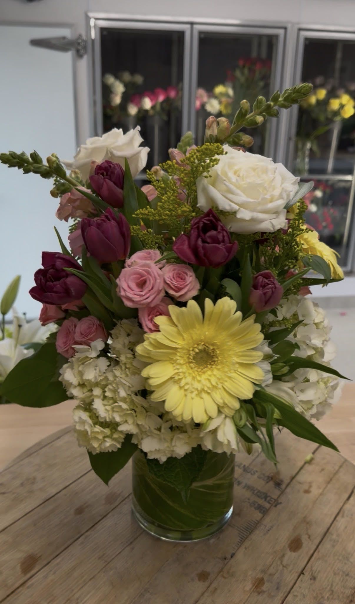 Mixed bouquet of yellow gerbera, white roses, pink spray roses, and dark tulips in a clear glass vase
