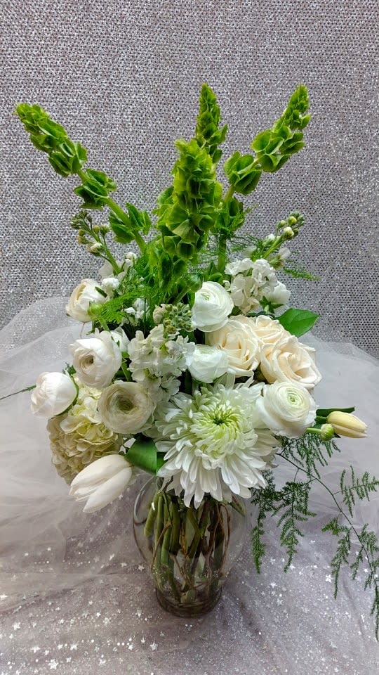 Tall white and green flower arrangement with roses, ranunculus, tulips, and Bells of Ireland in a clear glass vase
