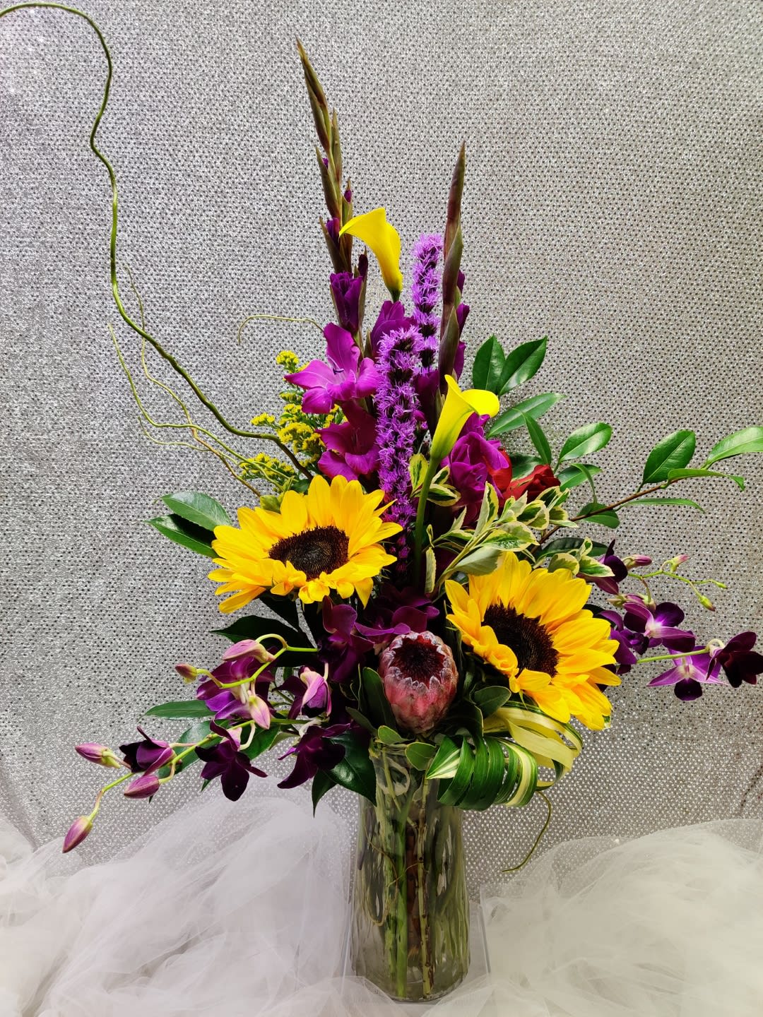 Tall mixed arrangement with sunflowers, purple orchids, yellow calla lilies, and protea in a clear glass vase.