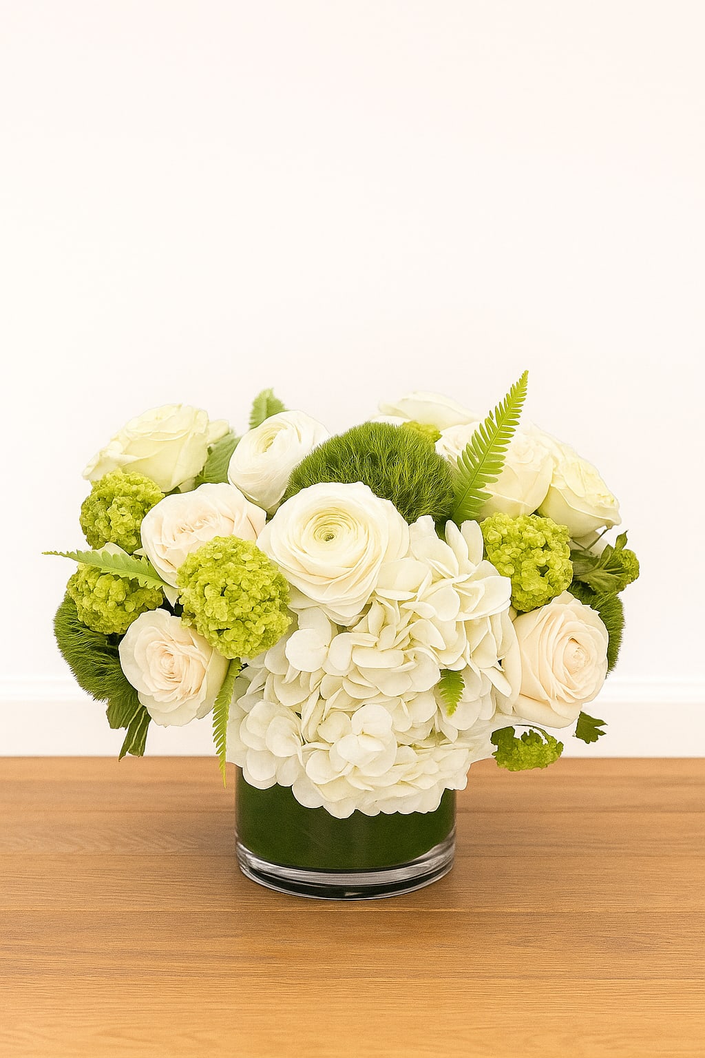 Low arrangement of white hydrangeas and cream roses with green accents in a clear glass cylinder vase