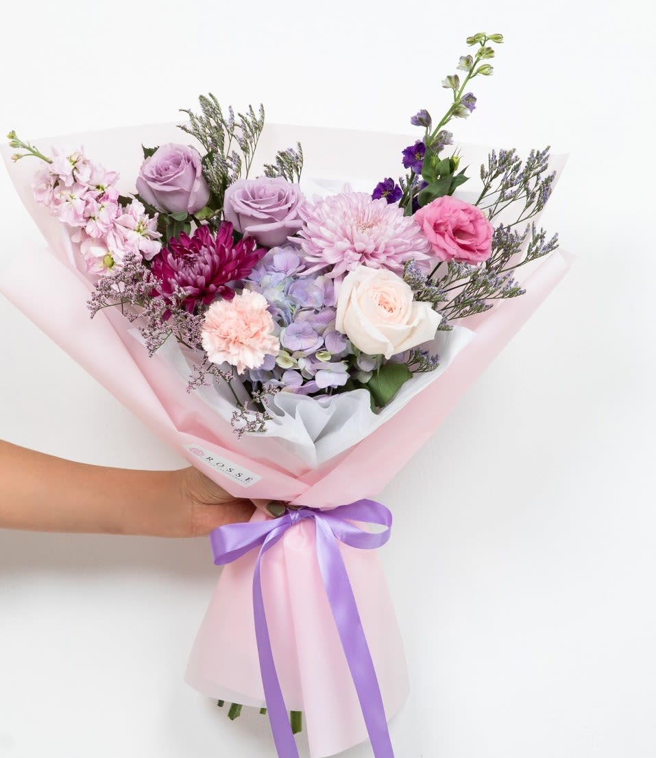 Lilac Dream  - A front-facing bouquet with different tones of lilac, lavender, and purple, wrapped in pink paper.   Flower types may vary depending on the season.  