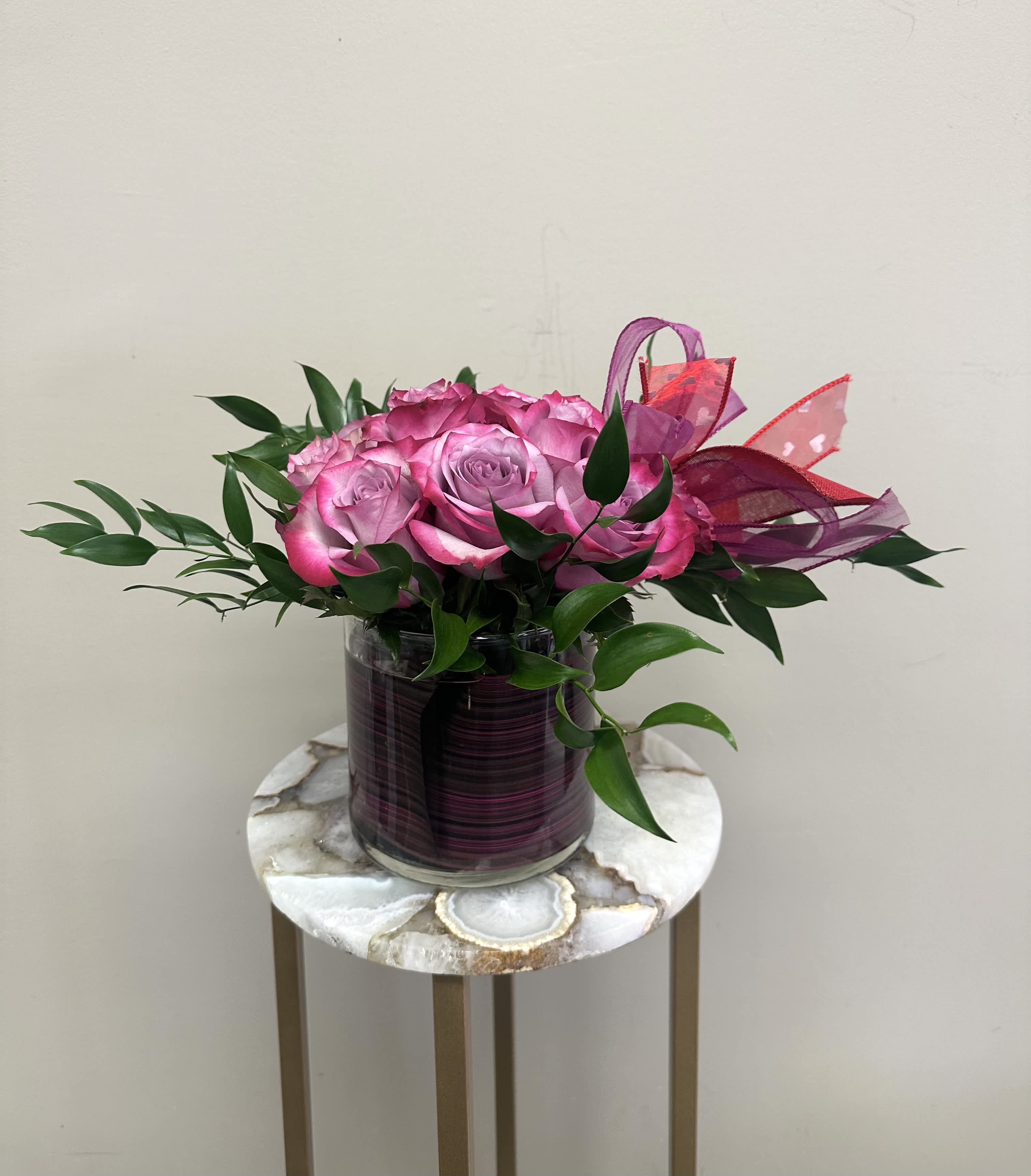 Low arrangement of bright pink roses in a glass cylinder vase with pink ribbon accents.
