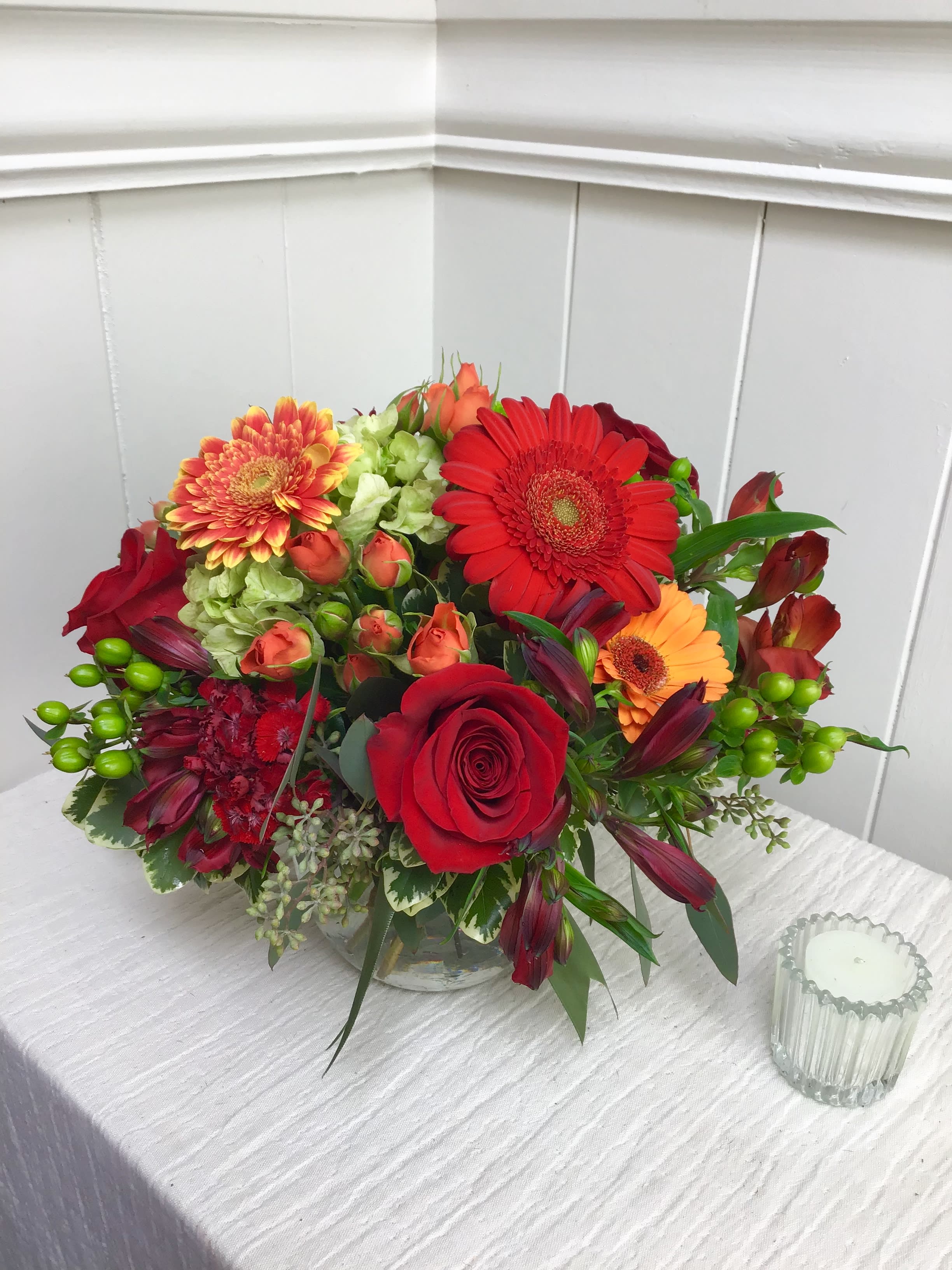Low round arrangement of red roses, gerbera daisies and mixed orange blooms in a clear glass bowl vase